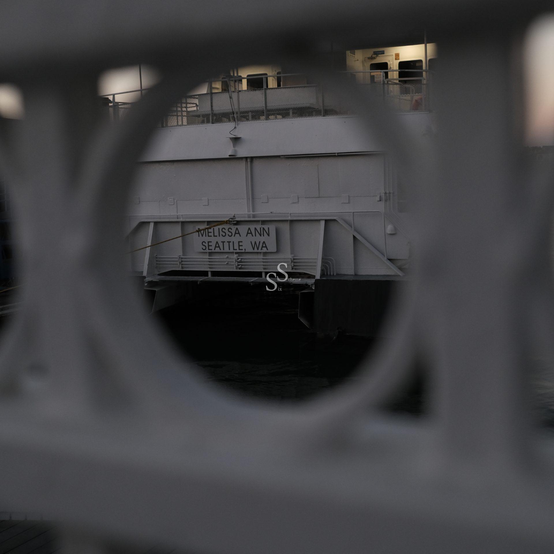 A white ferry named “Melissa Ann, Seattle, WA” is seen through a circular opening in a metal railing, with the water and part of the vessel visible in the background.