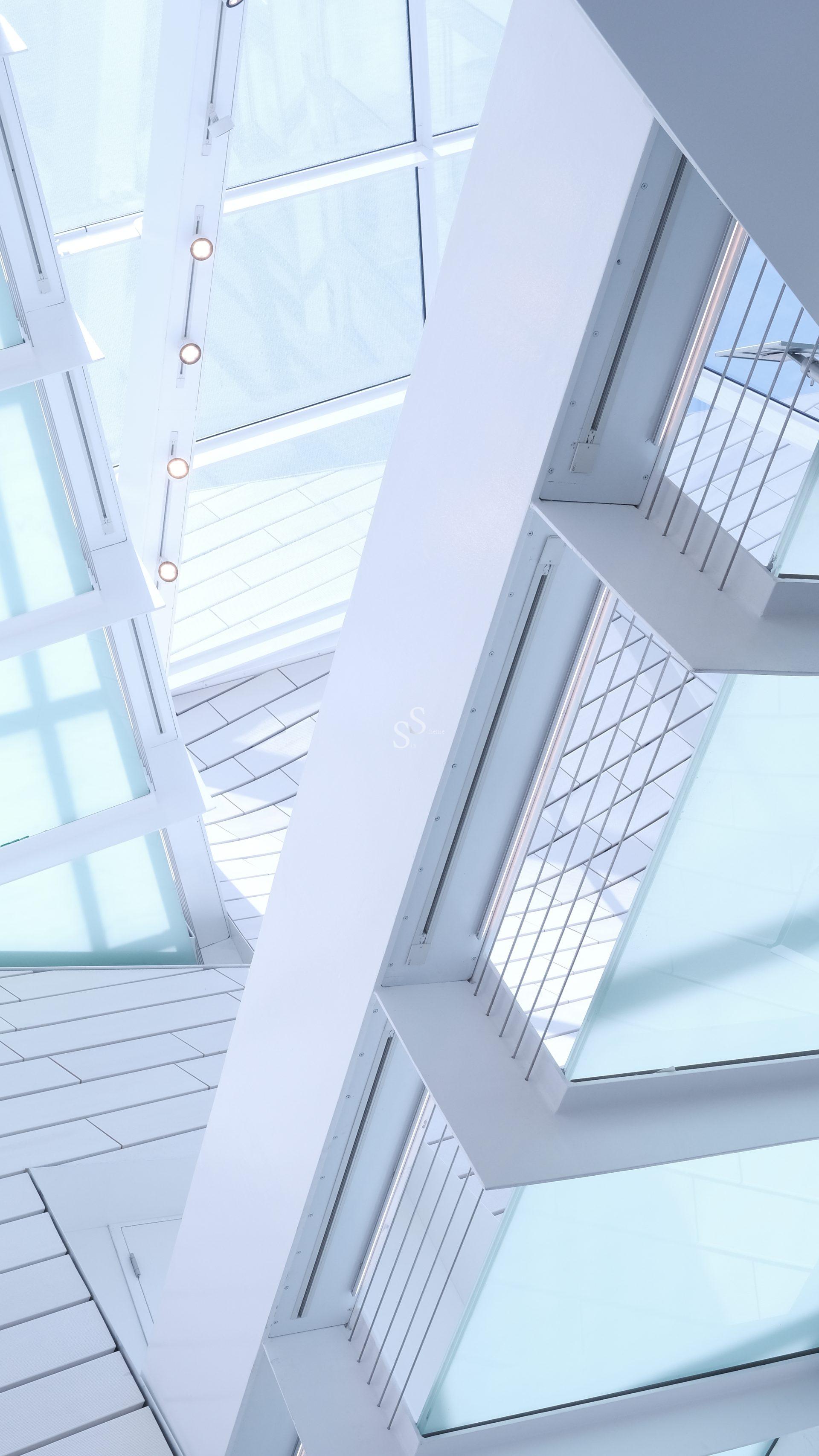 A modern architectural interior featuring white geometric beams, glass panels, and railings, with natural light streaming through large skylights overhead. The design is bright, minimalistic, and angular.
