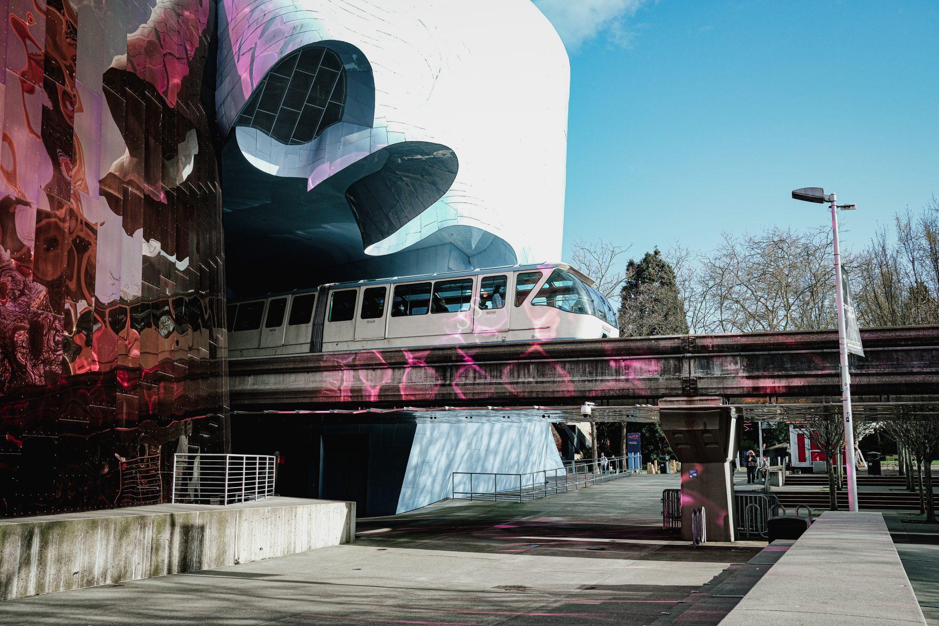 A white monorail train passes through a futuristic building with reflective, pink and silver surfaces on a sunny day; trees and a clear blue sky are in the background.