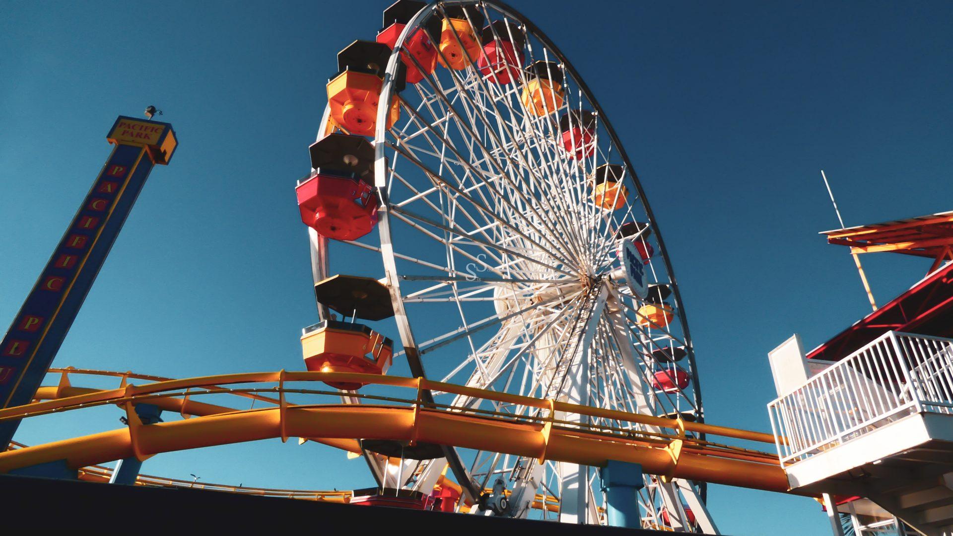 A large Ferris wheel with colorful red, yellow, and orange gondolas stands against a clear blue sky, next to a yellow roller coaster track at an amusement park.