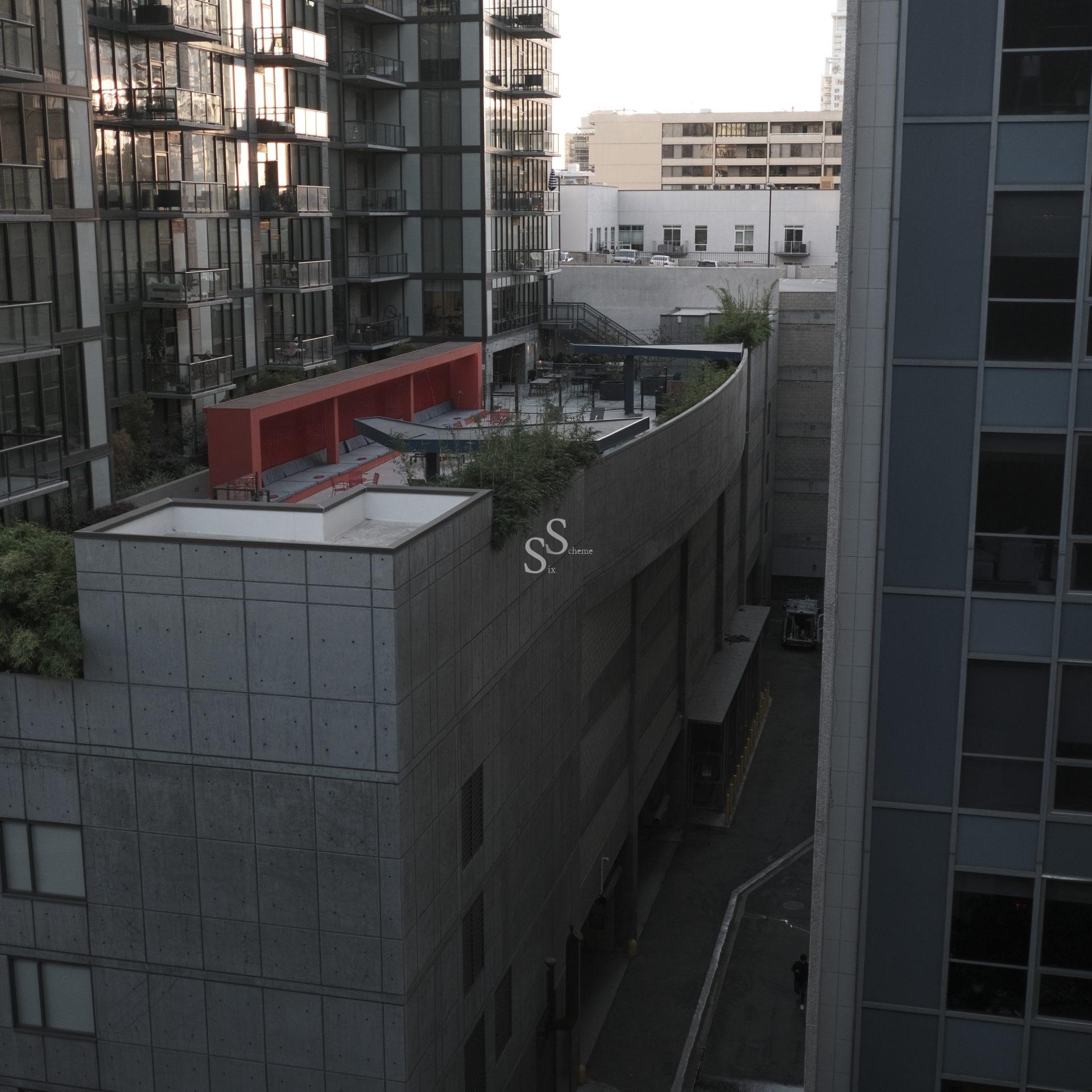 A narrow alley runs between tall, modern apartment buildings with glass facades; one building features a rooftop terrace with a red accent structure and some greenery.