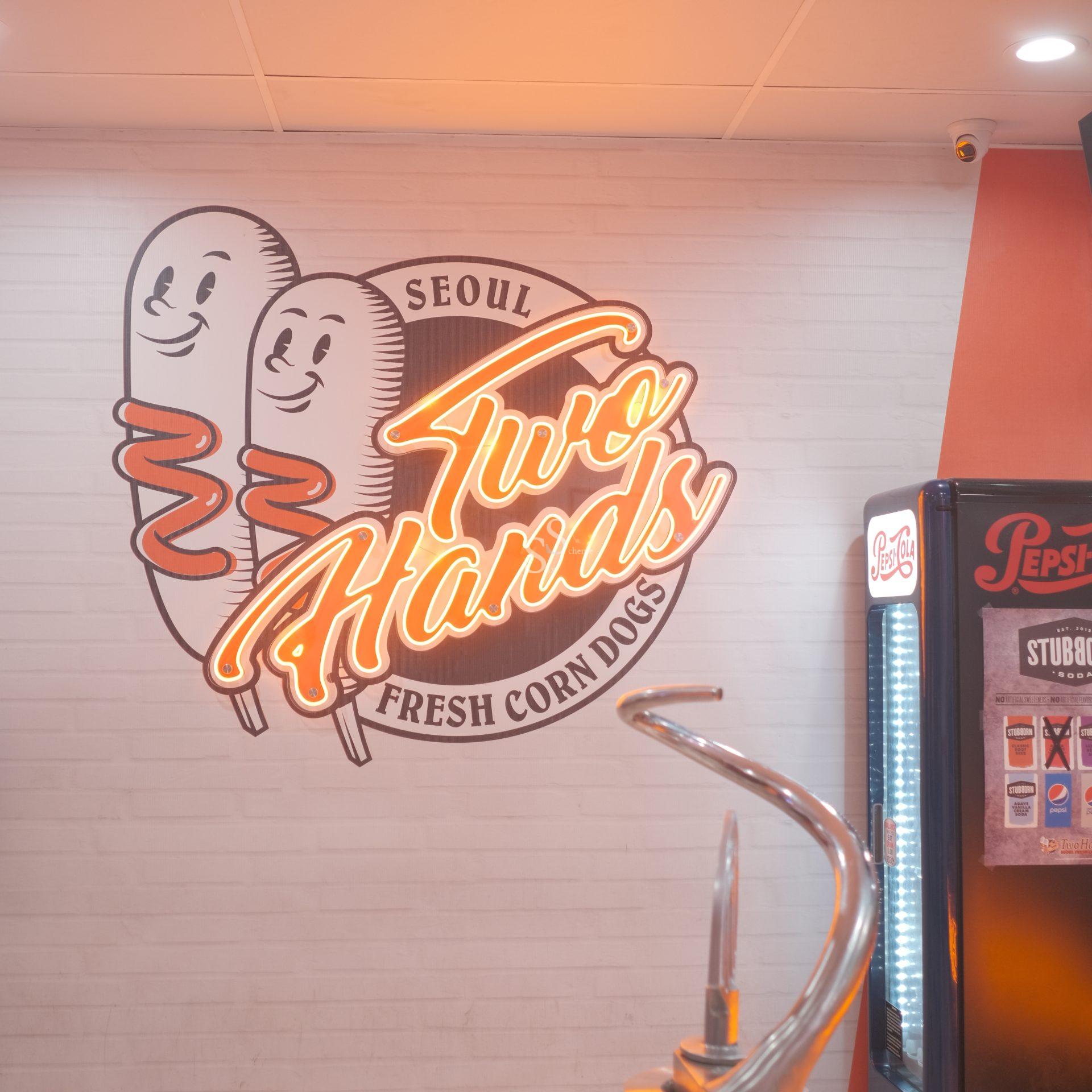 Wall with Two Hands Fresh Corn Dogs Seoul logo featuring two cartoon corn dogs. Nearby, part of a Pepsi fridge and a metal counter are visible. The setting appears to be a brightly lit restaurant.