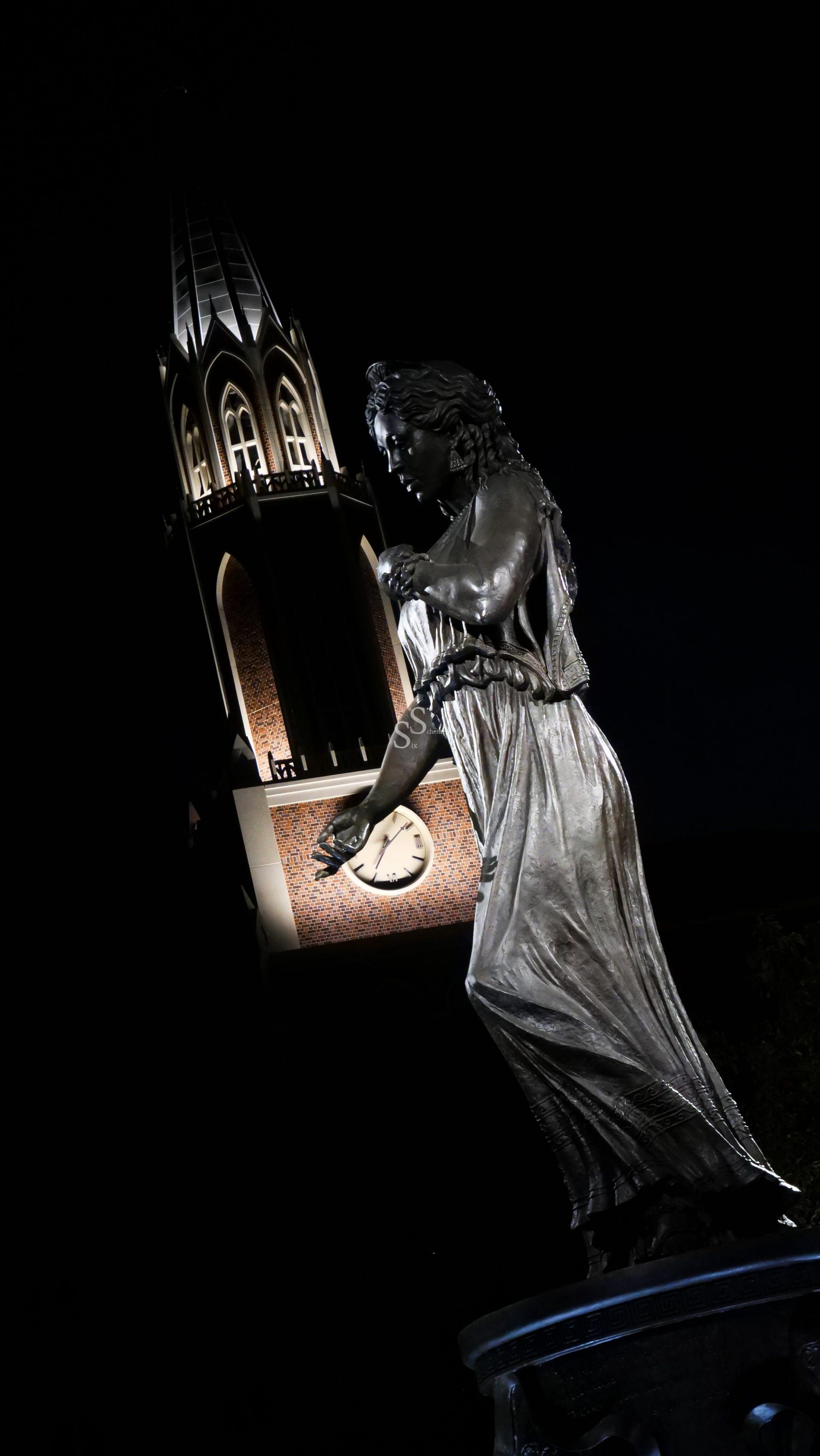 A bronze statue of a woman stands in the foreground, illuminated at night, with a tall, Gothic-style clock tower glowing in the background. The statue appears to be in motion, and the tower’s clock face is visible.
