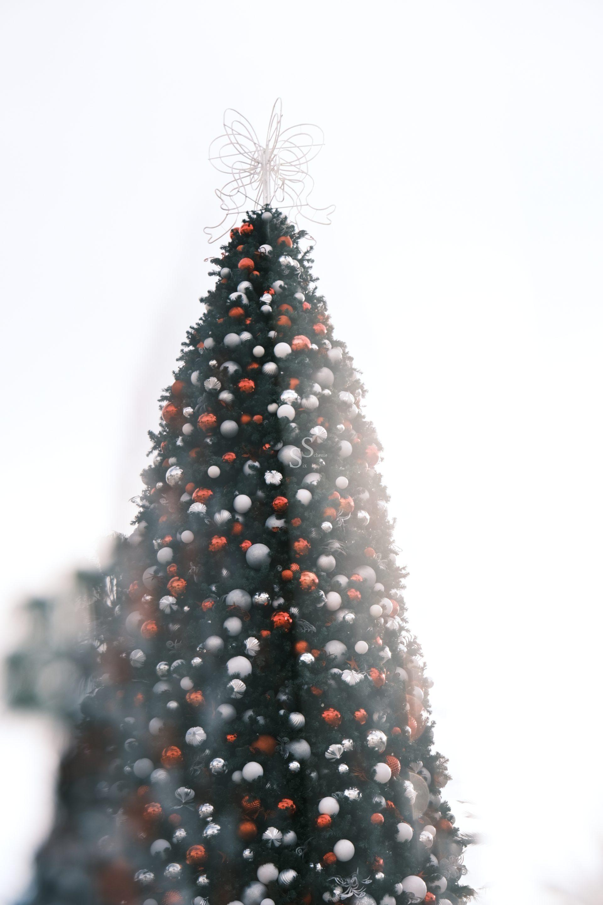 A tall Christmas tree decorated with red and white ornaments stands outdoors, topped with a wire angel. The background is bright and slightly hazy, creating a festive atmosphere.