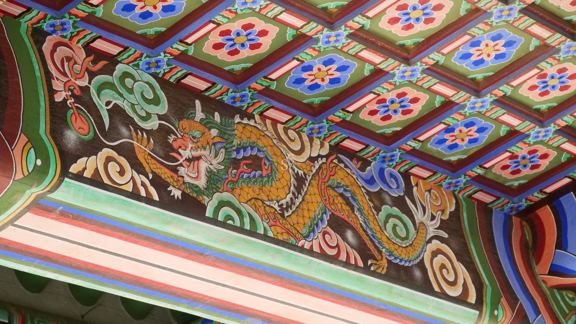 Colorful ceiling detail in a traditional Korean building, featuring a painted yellow dragon with green, red, and blue accents, surrounded by floral patterns and geometric designs.