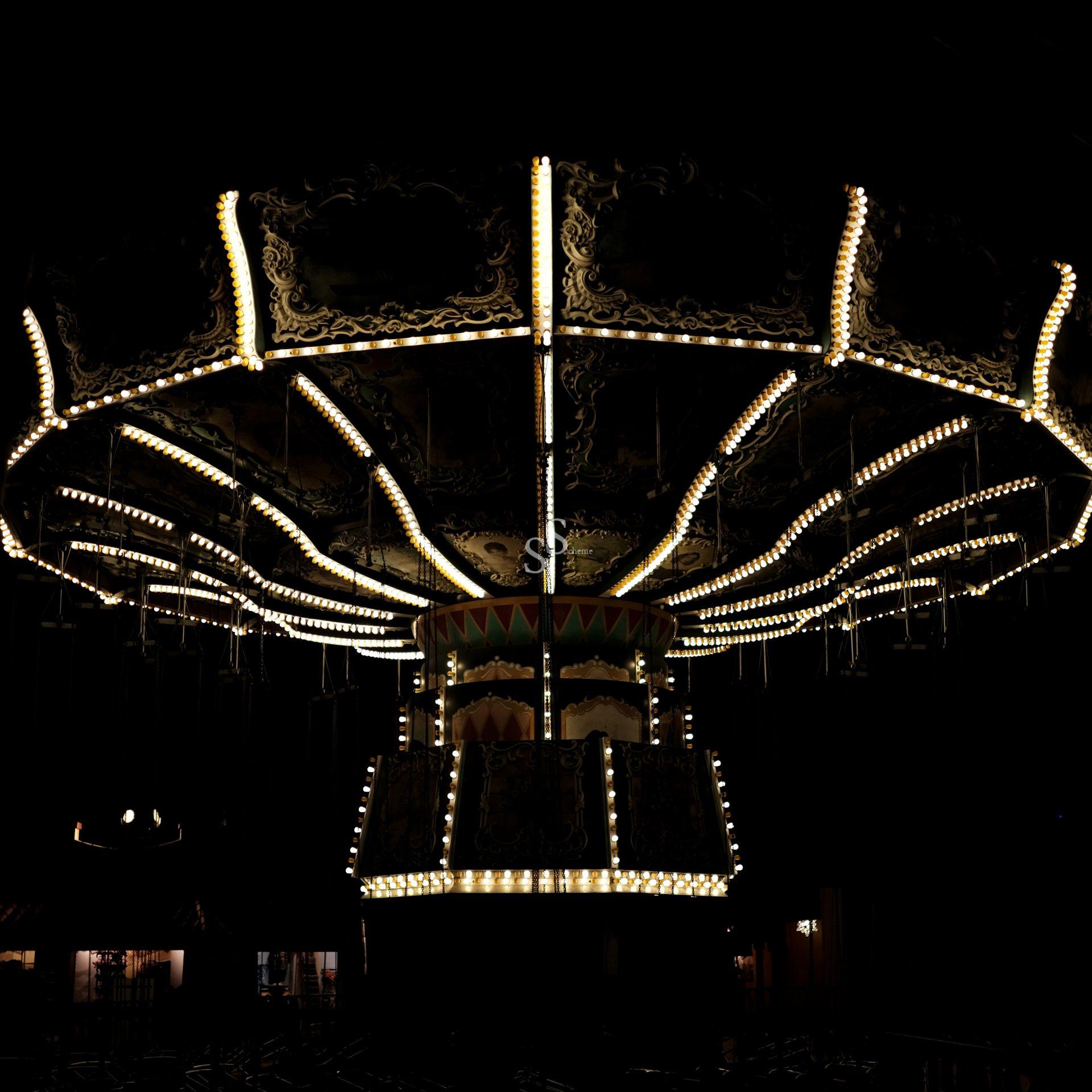 Illuminated brightly at night, a carousel swing ride's ornate details and empty hanging chains stand out against the dark background.