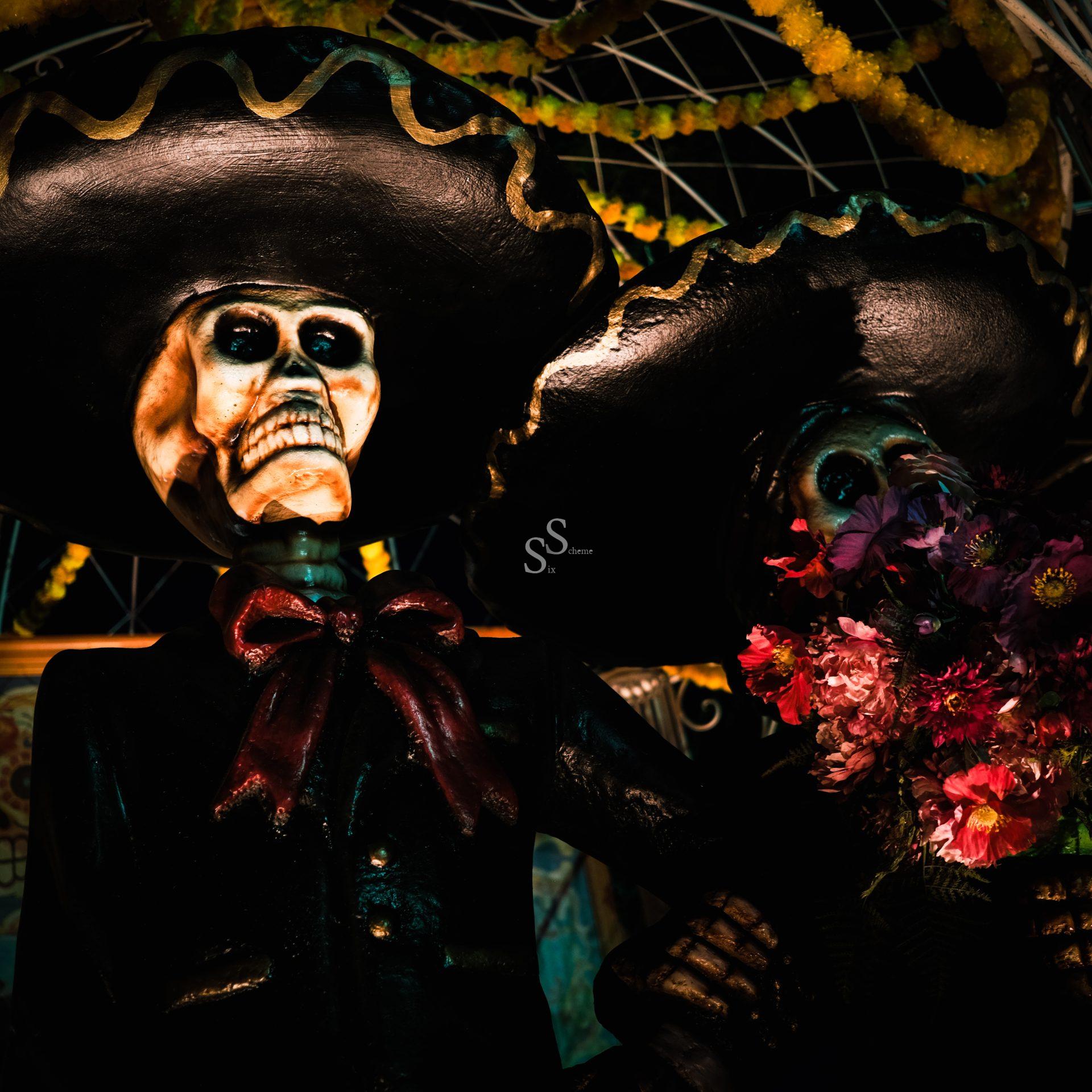 Two large skeleton figures wearing black sombreros and suits are shown. One holds a bouquet of flowers. The scene is decorated with colorful garlands, evoking a Day of the Dead celebration.