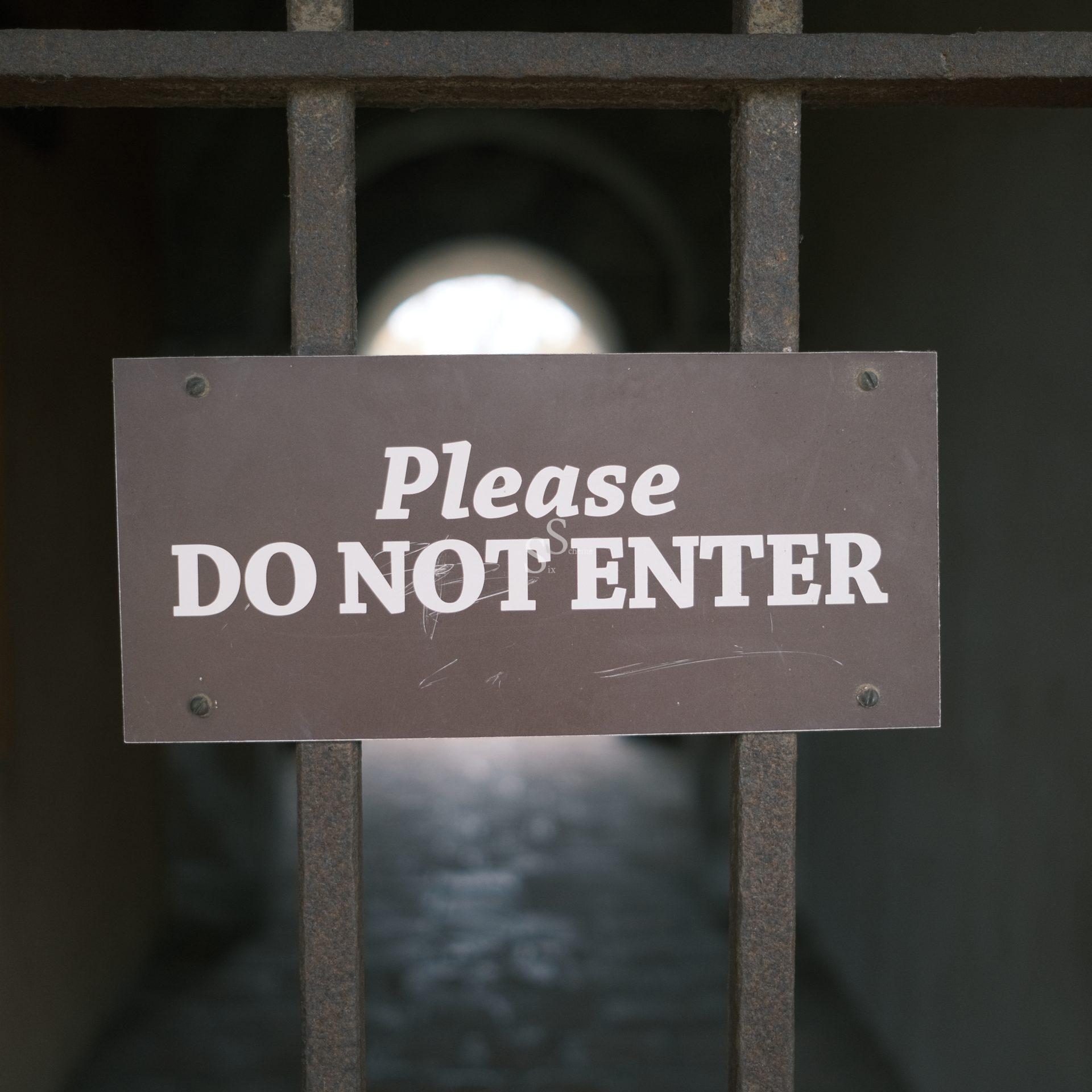 A metal gate with a sign that reads Please DO NOT ENTER blocks the entrance to a dimly lit passageway. Light is visible at the far end of the tunnel.