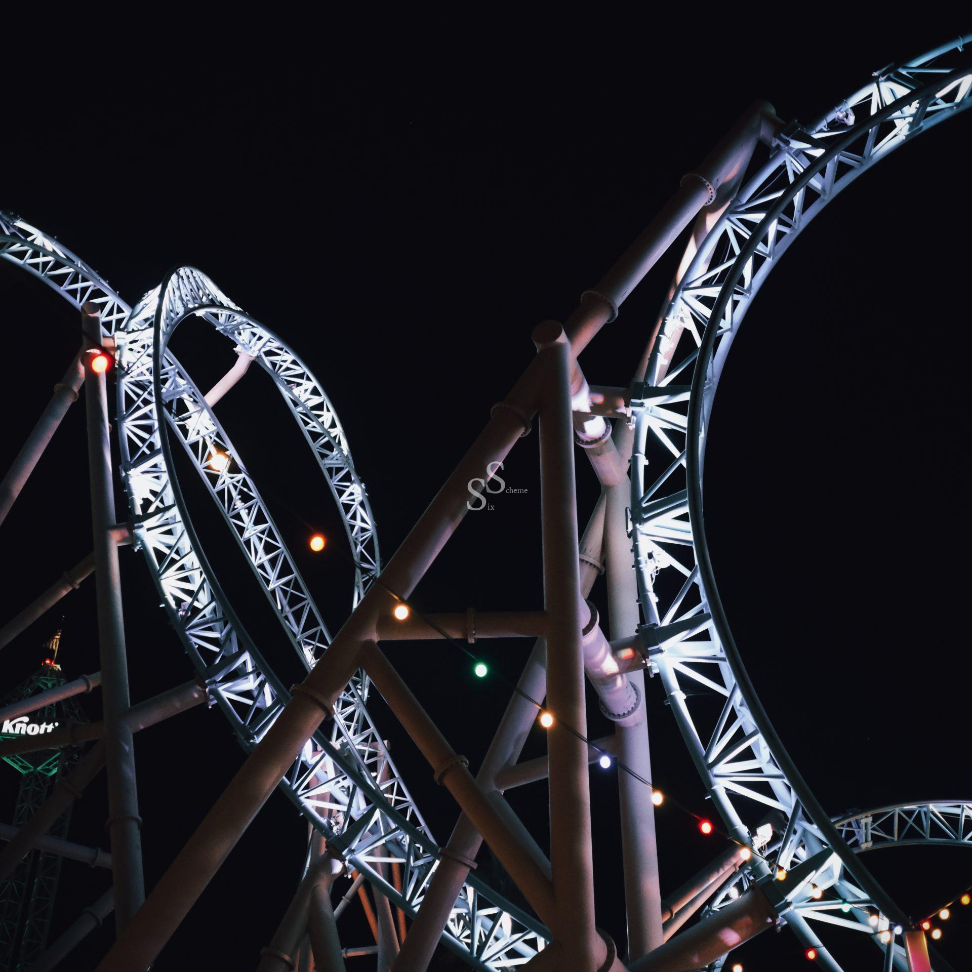 A roller coaster with bright white metal loops is illuminated at night. Multicolored string lights hang across the lower part of the image, contrasting against the dark sky.