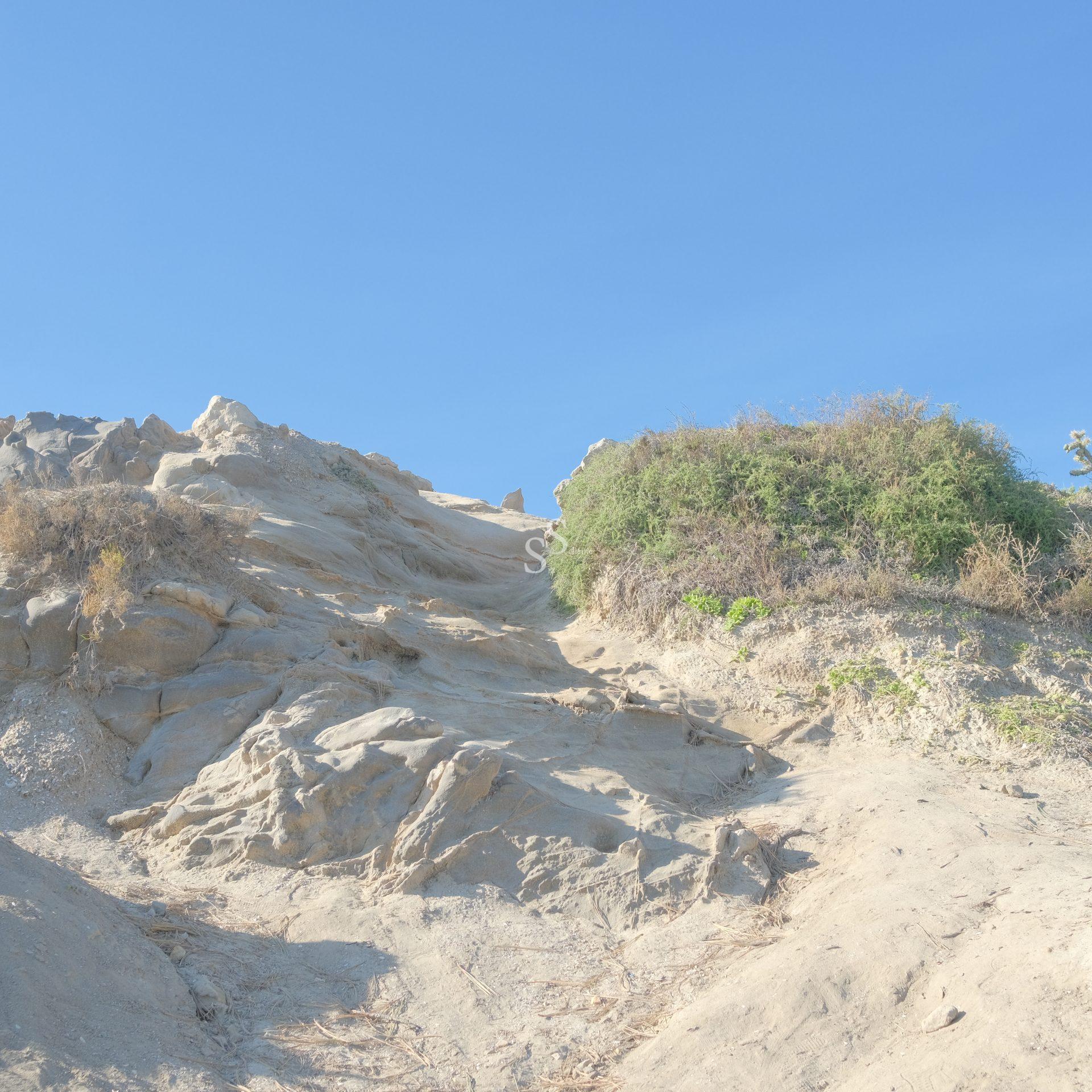 A rocky, uneven hillside with patches of dry grass and green shrubs under a clear blue sky, illuminated by bright sunlight.