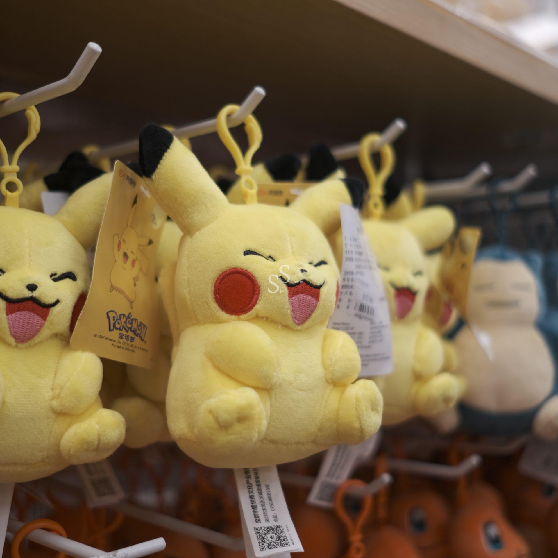A display rack in a store features hanging, smiling Pikachu plush keychains, backed by other soft toys.