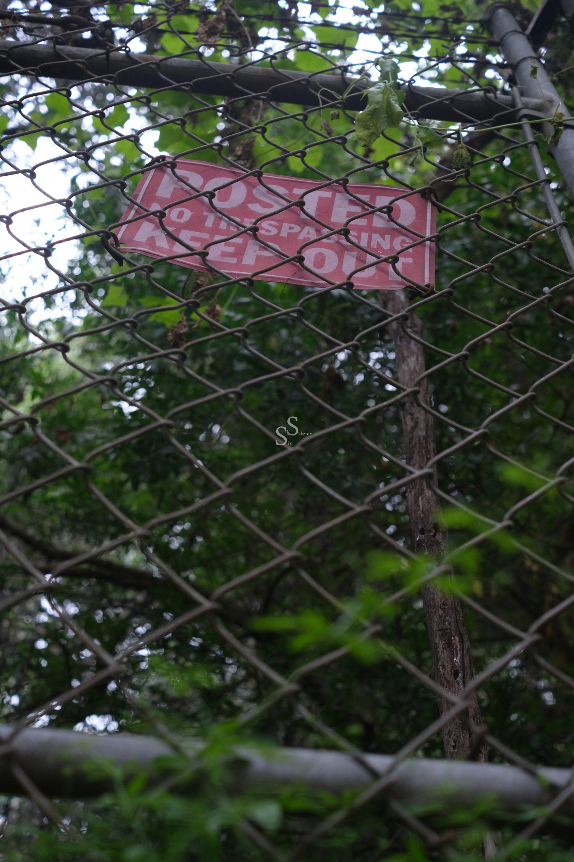 A weathered POSTED NO TRESPASSING KEEP OUT sign is attached to a chain-link fence, surrounded by overgrown greenery and trees in the background.