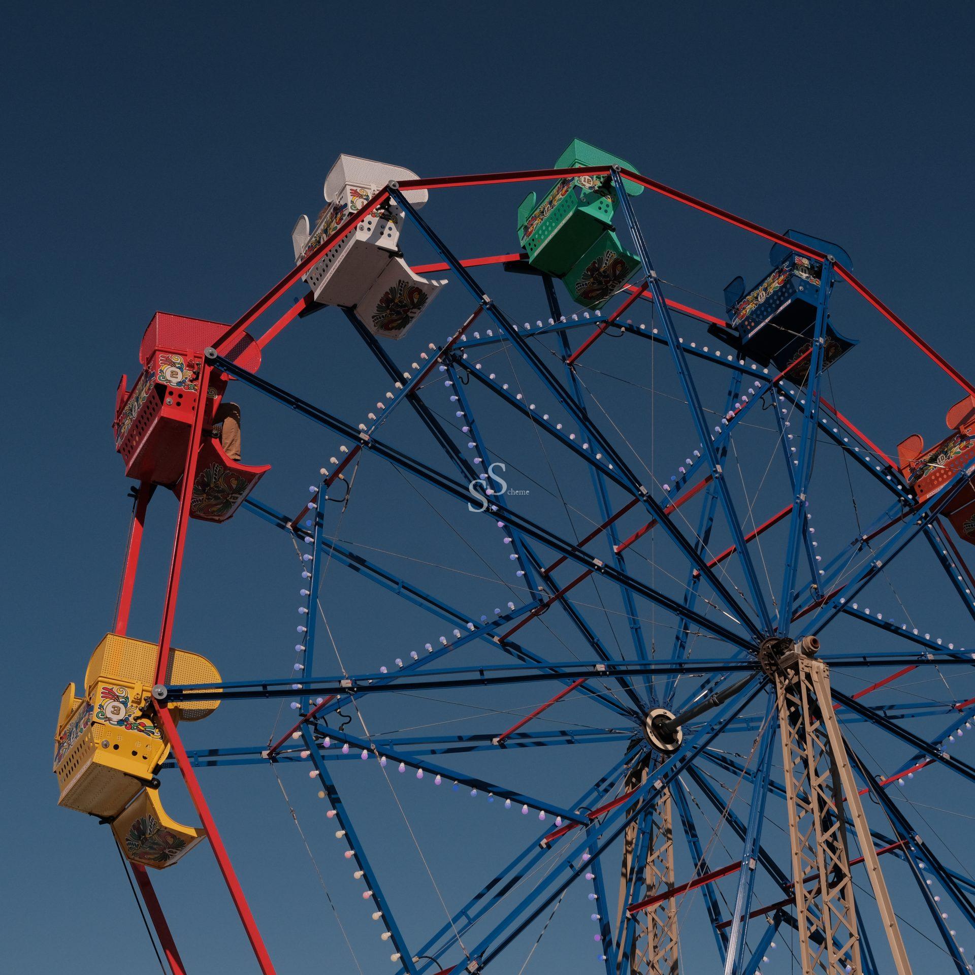 A colorful Ferris wheel with red, yellow, white, green, and blue gondolas stands against a clear blue sky, seen from a low angle. The structure is made of blue and red metal beams.