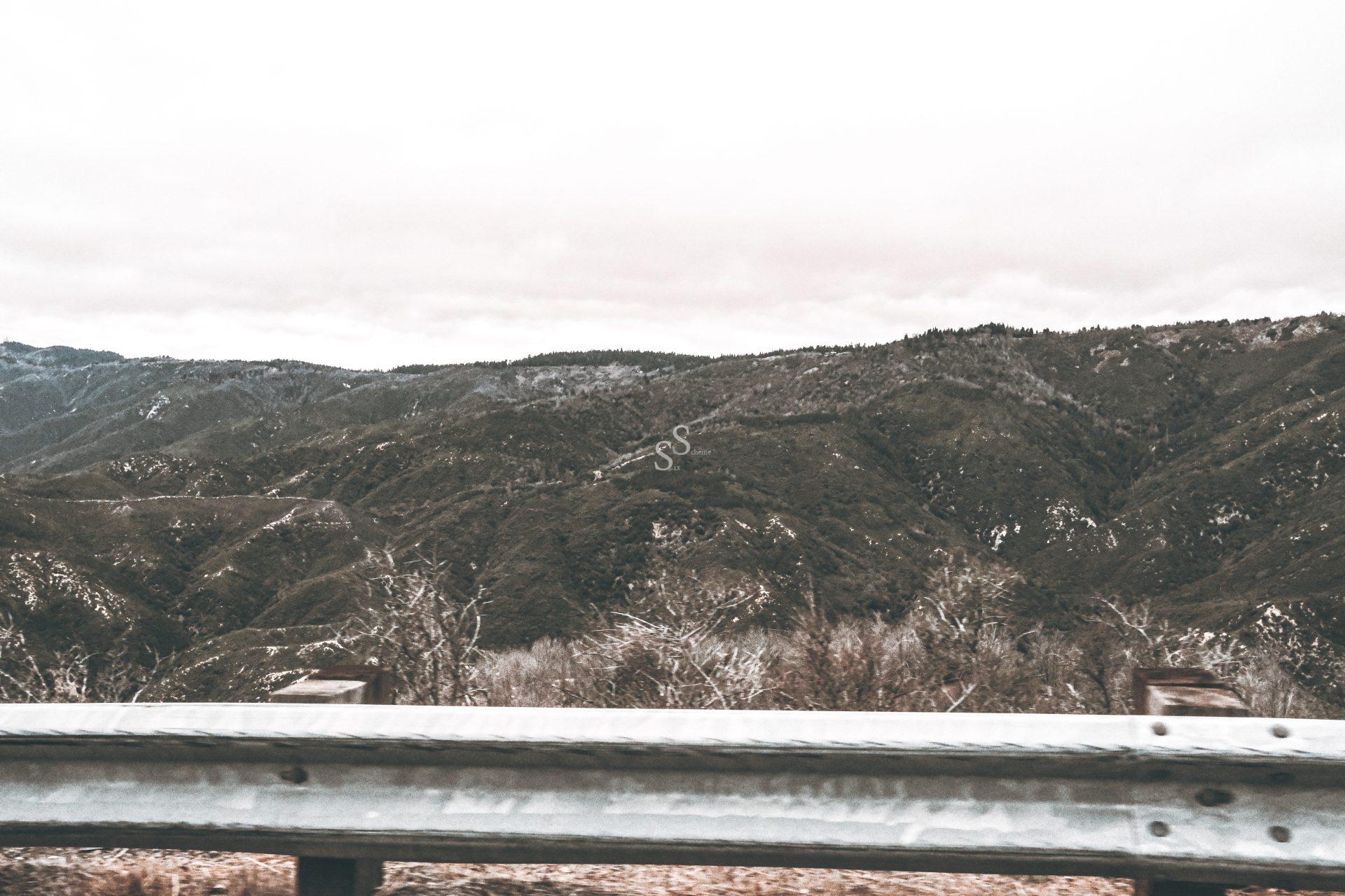 A metal guardrail lines the edge of a road, overlooking rolling green hills and dense forests under a cloudy sky. The landscape appears rugged and mountainous, with a muted, overcast atmosphere.