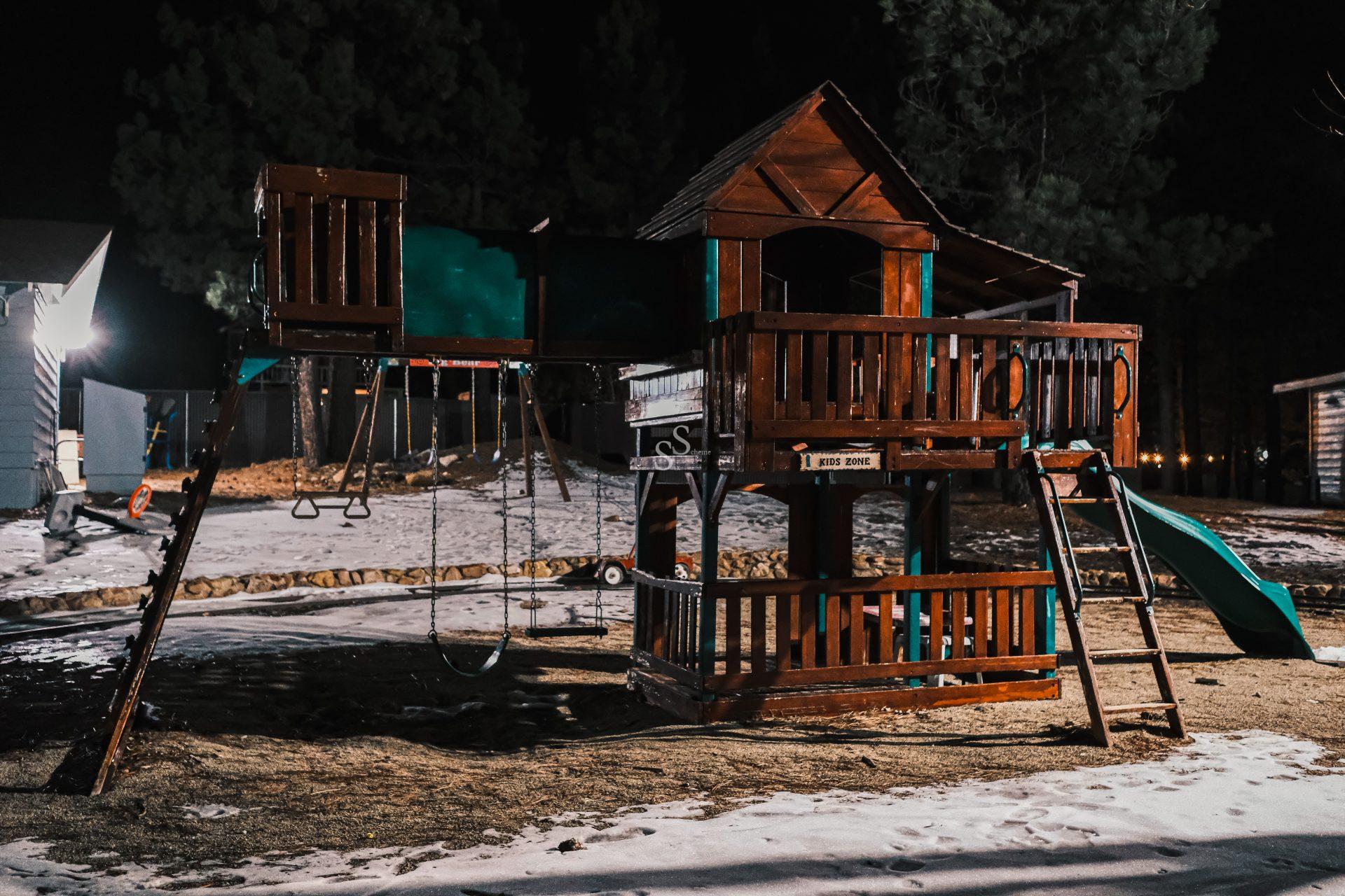 A wooden playground set at night features swings, a slide, and a small clubhouse, surrounded by patches of snow and lit by artificial lights. Trees and buildings are visible in the background.