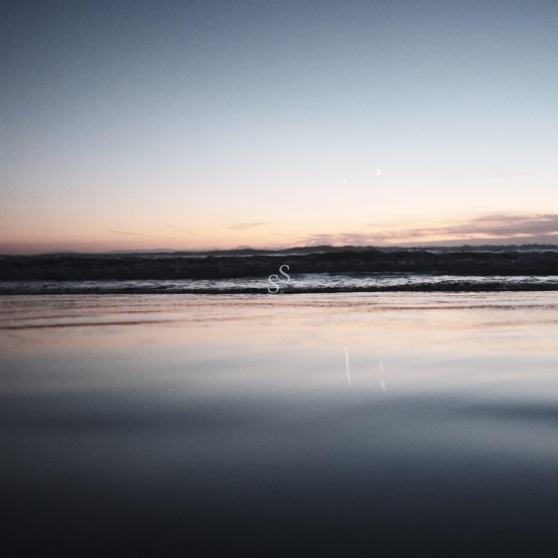 A calm ocean at sunset with soft waves and a clear sky fading from pale orange near the horizon to blue, reflecting gentle light on the water’s surface.