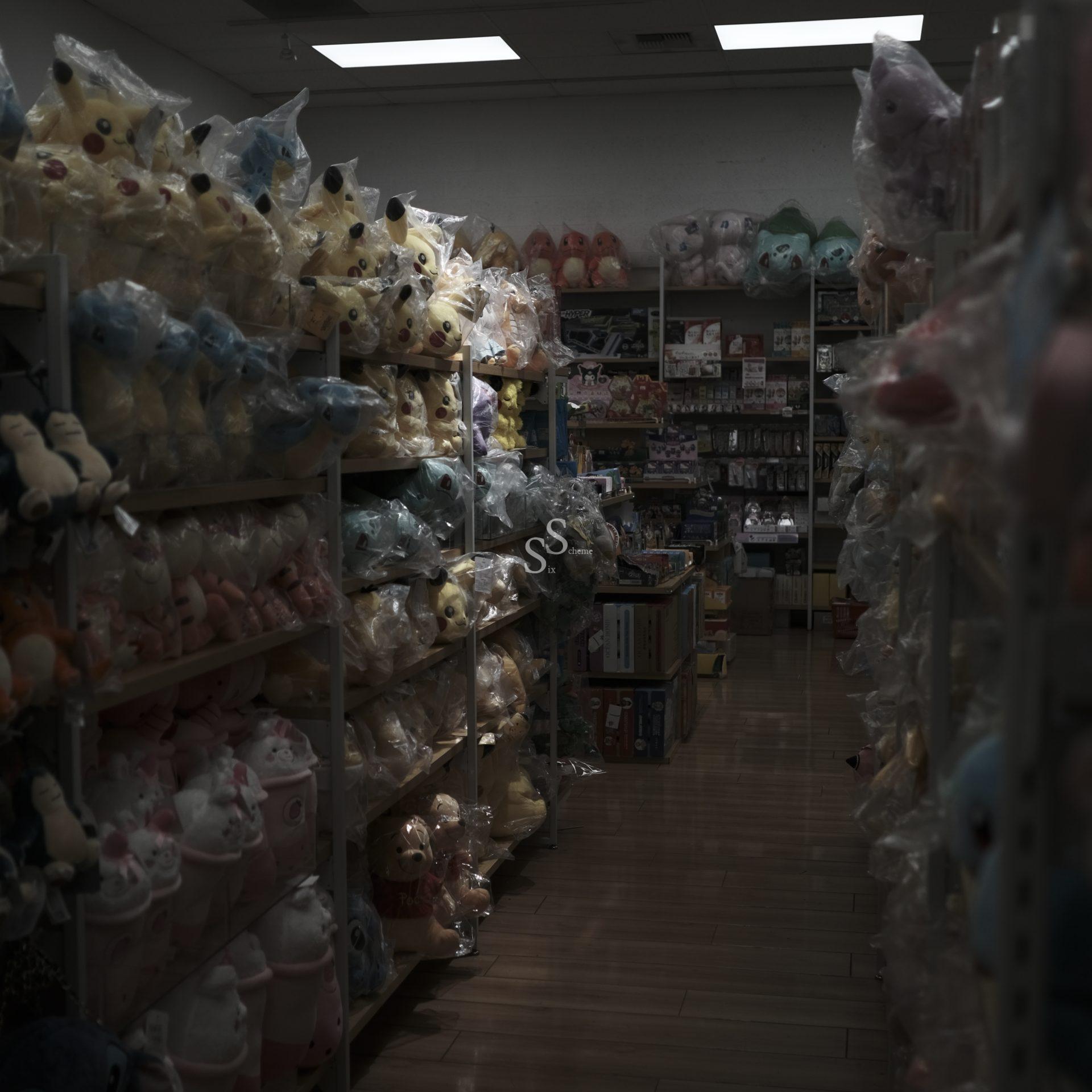 Dimly lit store aisle filled with shelves stocked with plush toys in plastic bags, featuring many yellow Pikachu dolls and other colorful characters, with more toys visible toward the back of the store.