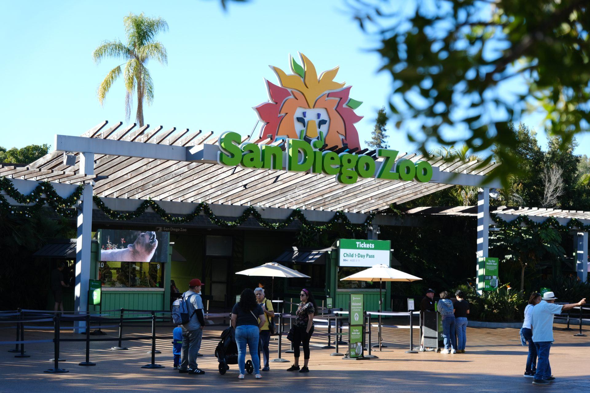 Entrance to the San Diego Zoo with a large sign featuring a lion logo, people walking toward ticket booths, greenery in the background, and a sunny sky overhead.