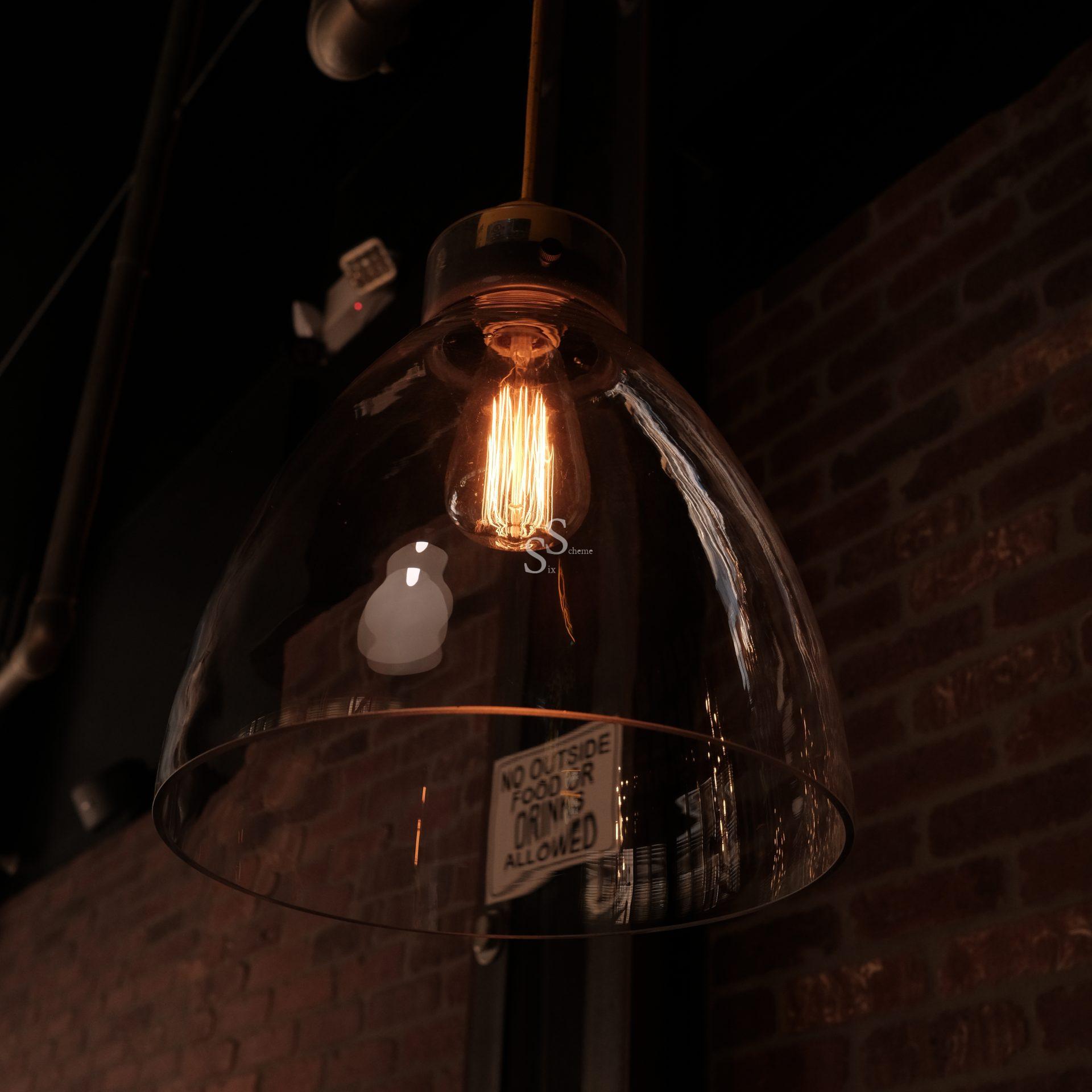 A clear glass pendant light with an exposed filament bulb hangs from the ceiling. A sign inside the lamp reads, “NO OUTSIDE FOOD OR DRINKS ALLOWED.” Brick wall and ceiling pipes are visible in the background.