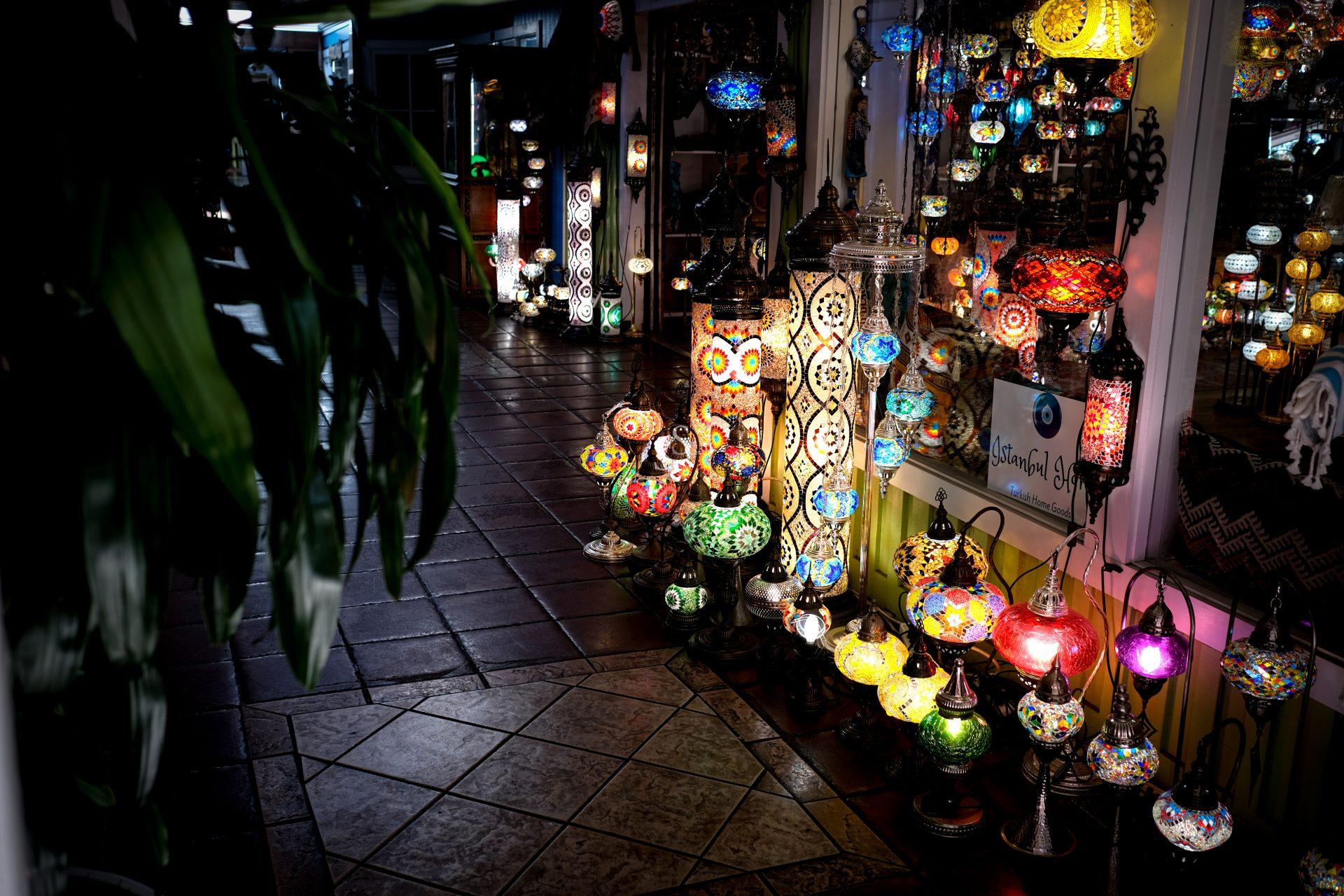 A variety of colorful, mosaic-patterned Turkish lamps are displayed on the floor and in windows of a dimly lit shop, casting vibrant light on the tiled floor.