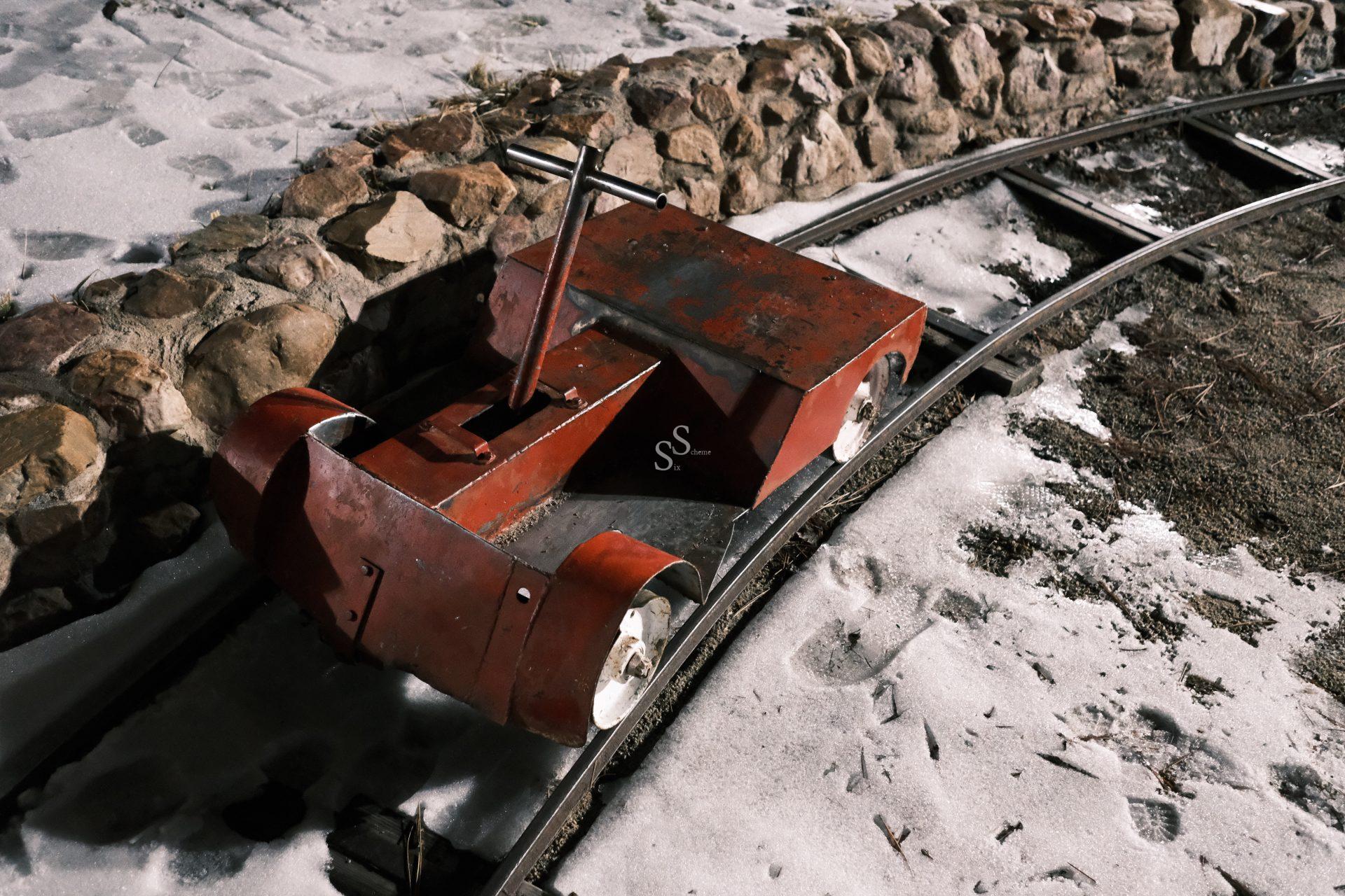 A small, red, metal handcar sits on narrow-gauge railway tracks surrounded by snow and gravel, with a stone wall lining the edge of the track.