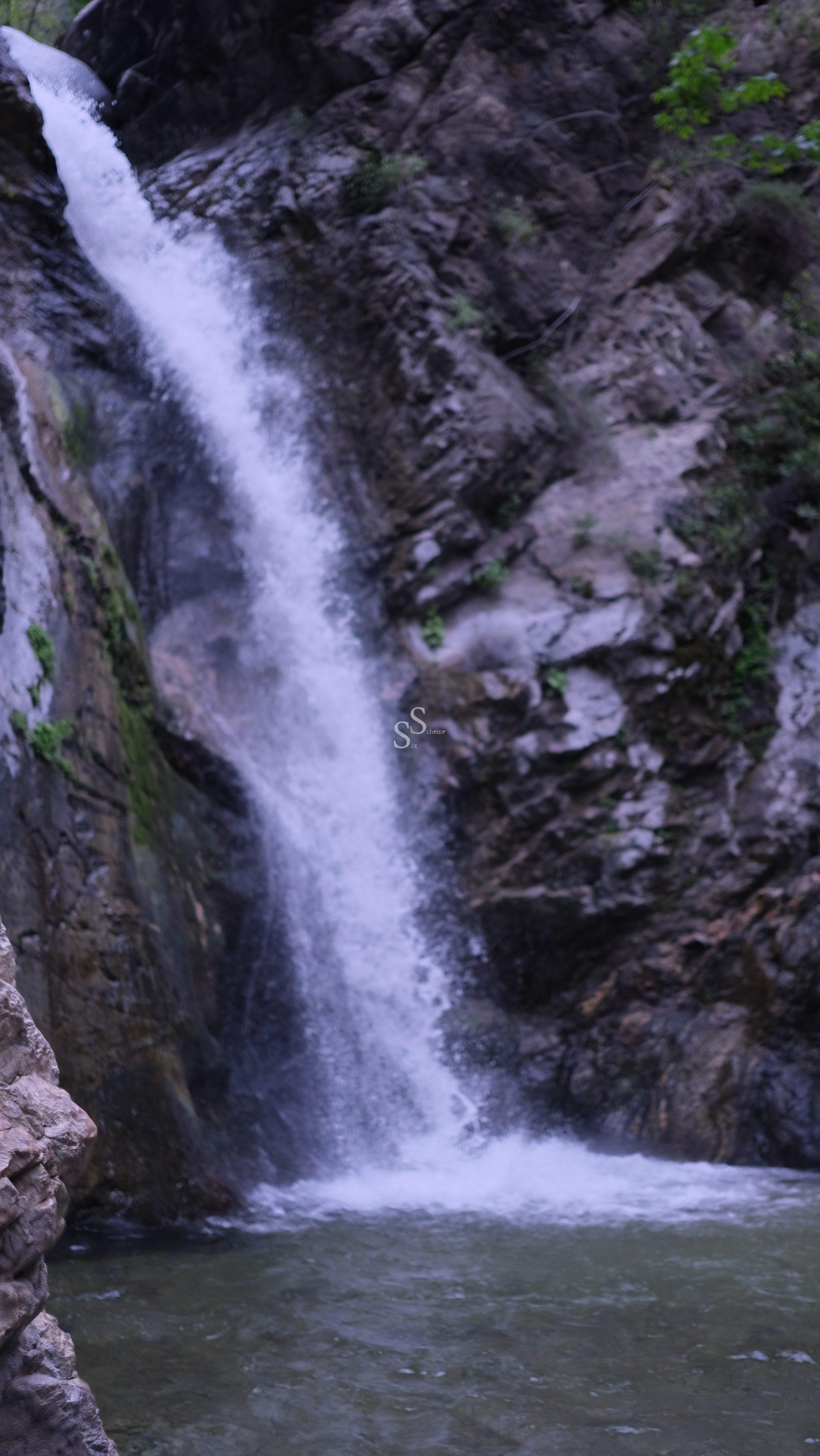 A waterfall cascades down a rocky cliff surrounded by moss and greenery, splashing into a small pool below. The natural scene is serene and features rugged rock formations.
