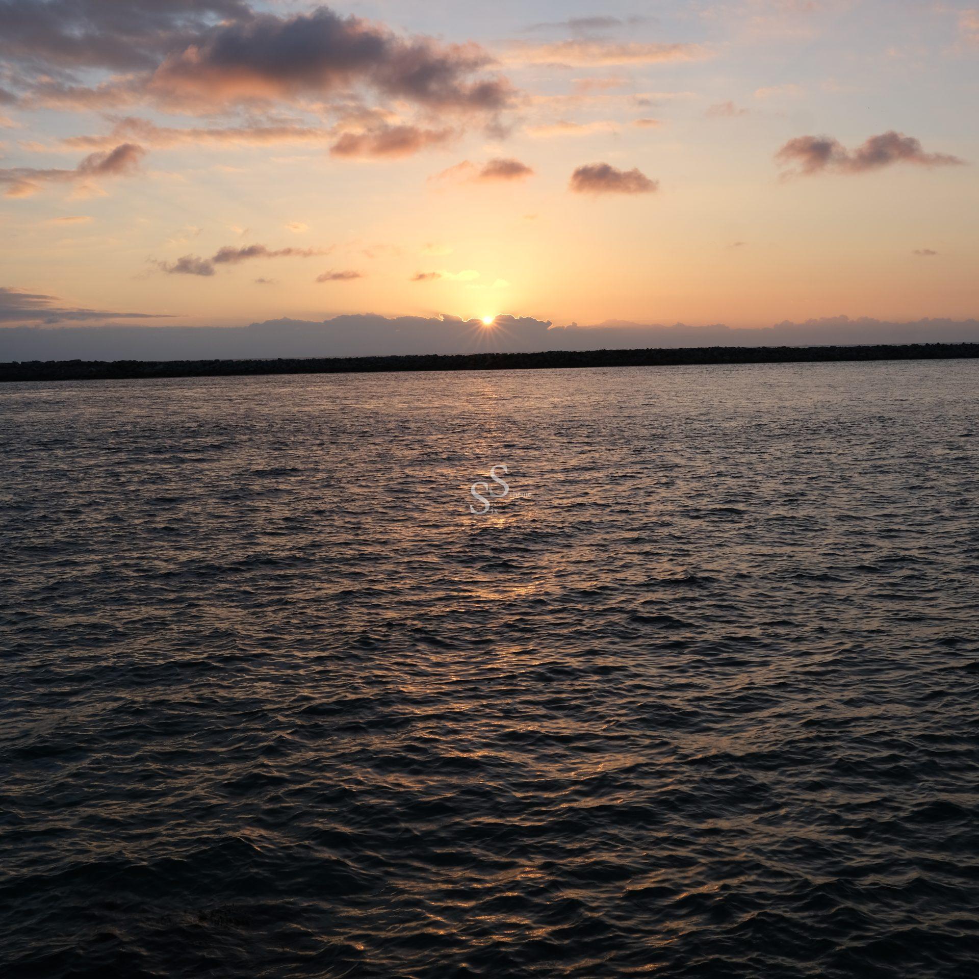 The sun sets behind distant clouds over a calm body of water, casting warm orange and pink hues across the sky and reflecting softly on the rippling surface.