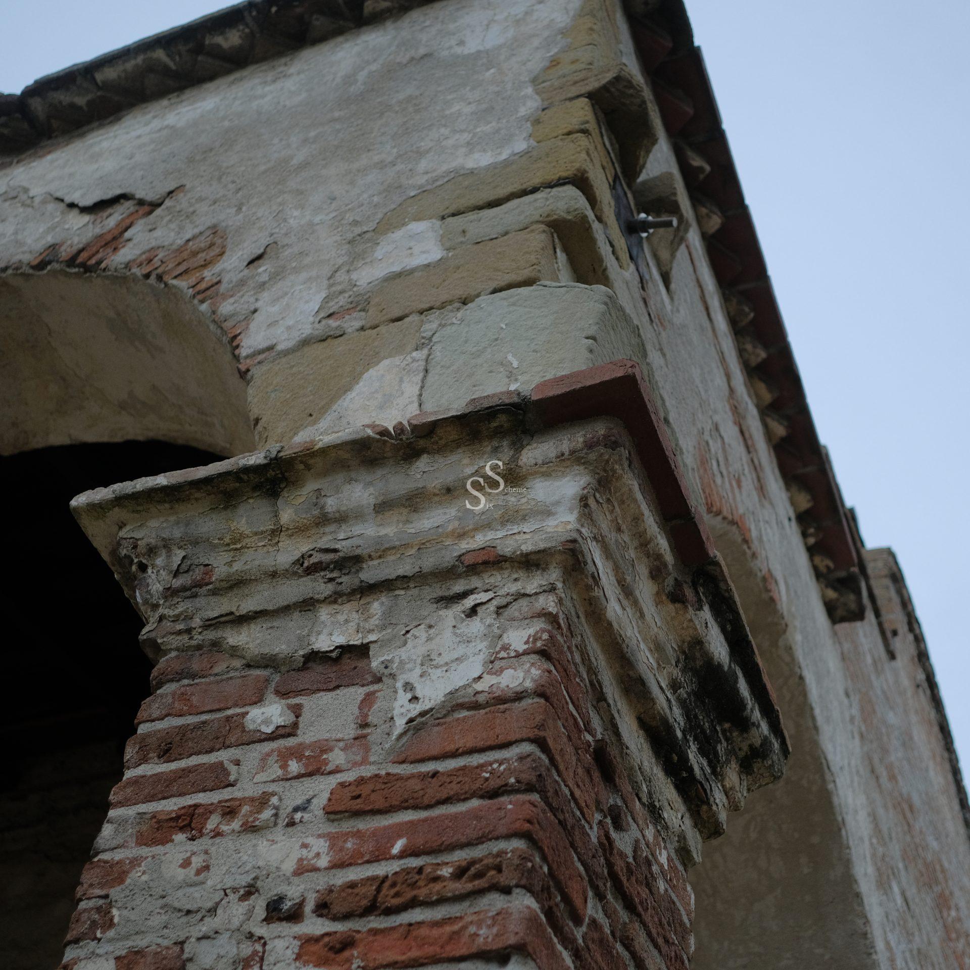 Close-up view of a weathered brick and stone building corner, showing peeling plaster, exposed bricks, and signs of age, set against a cloudy sky.