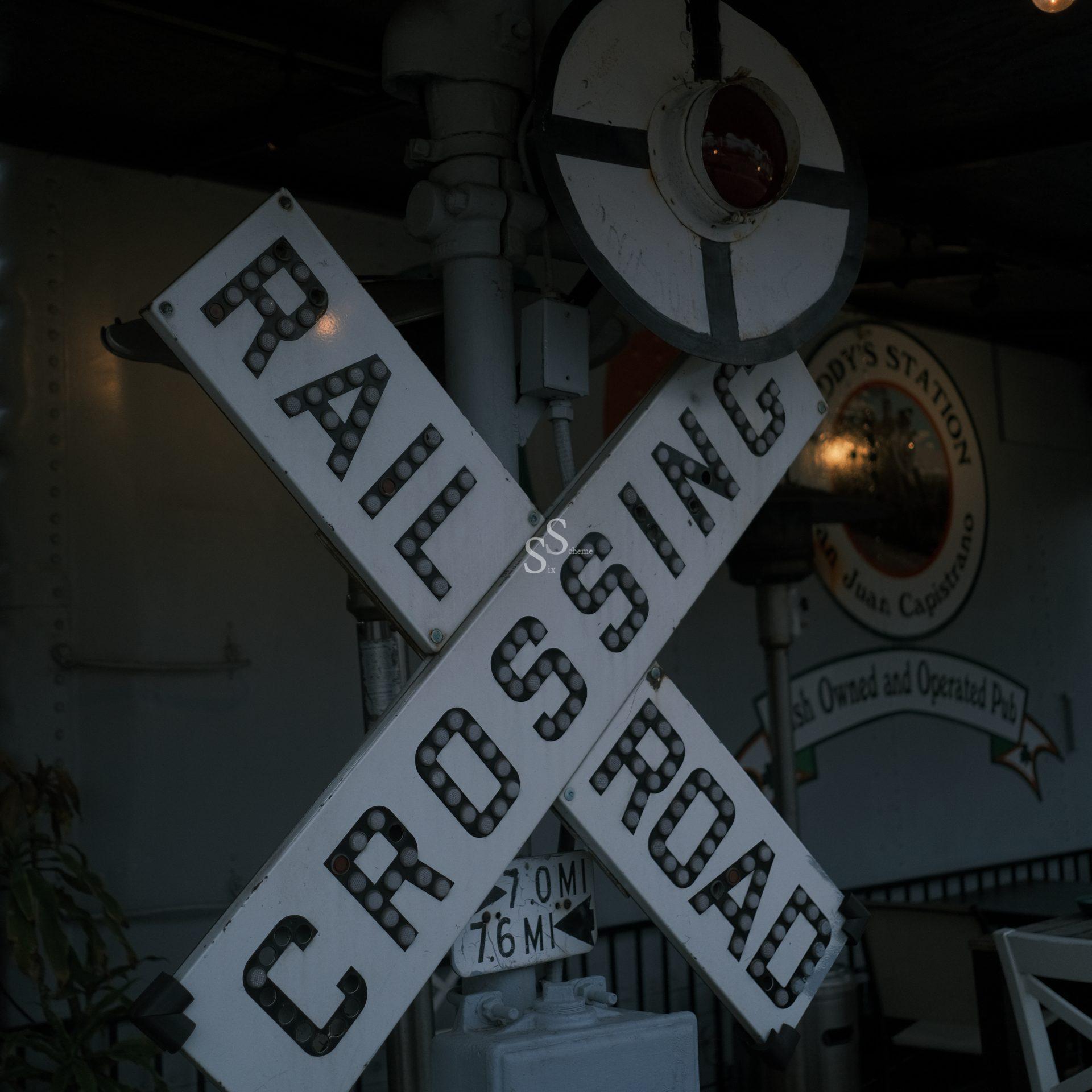 A vintage railroad crossing sign with bold letters stands in front of a wall bearing a partially visible circular sign that reads “Station.” The scene has a nostalgic, old-fashioned atmosphere.