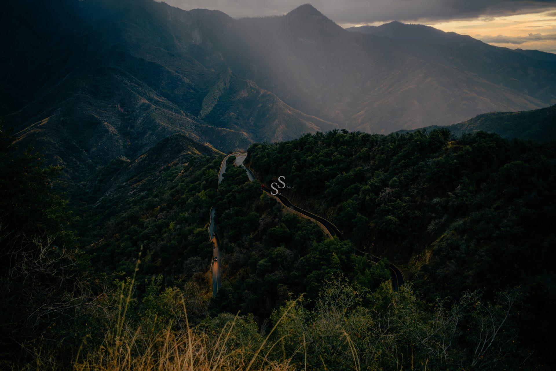 Winding mountain road cutting through dense green forest with tall peaks in the background, illuminated by rays of sunlight breaking through a cloudy sky.