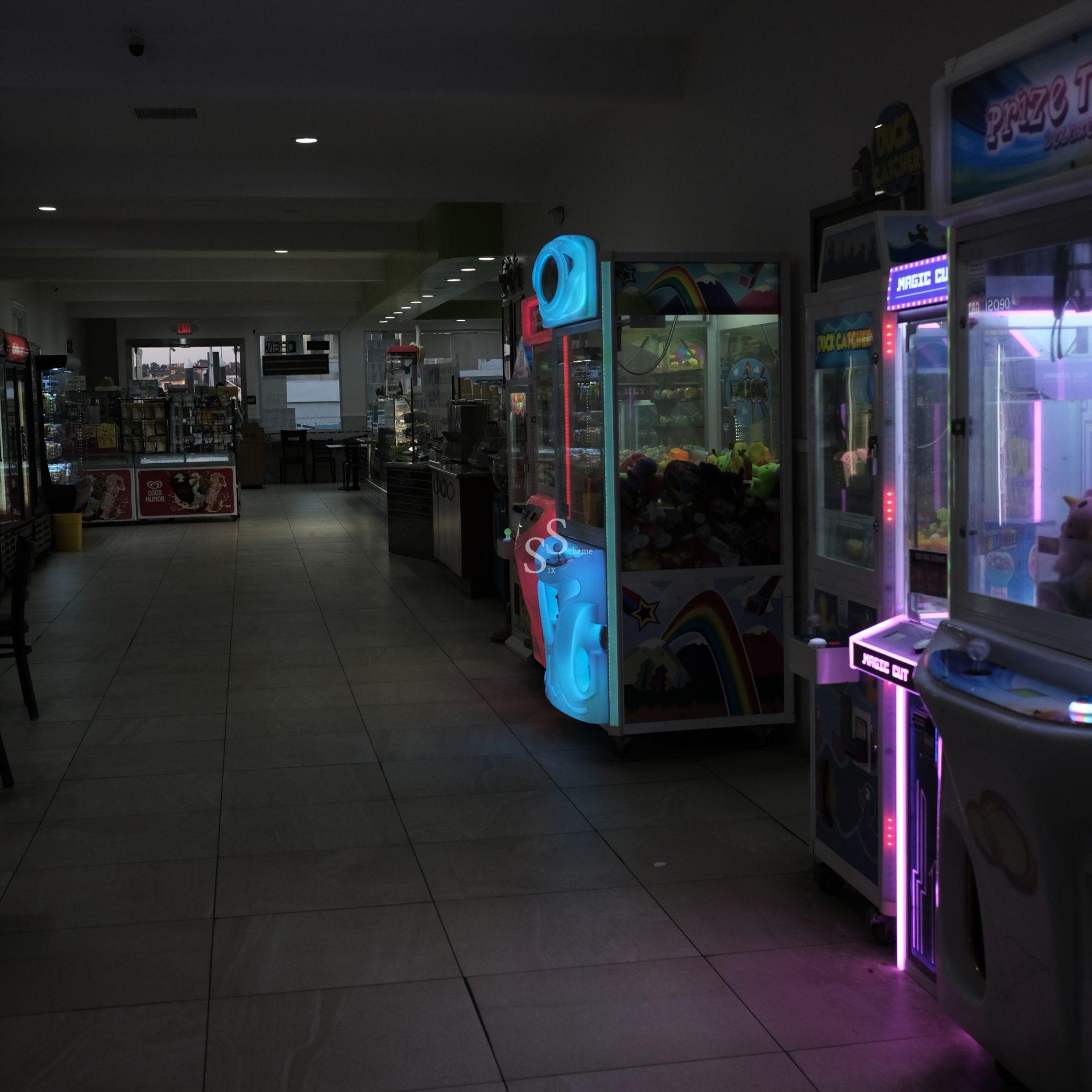 A dimly lit arcade area with claw machines glowing in blue and pink lights on the right, and a mostly empty food court with tables and chairs visible in the background.
