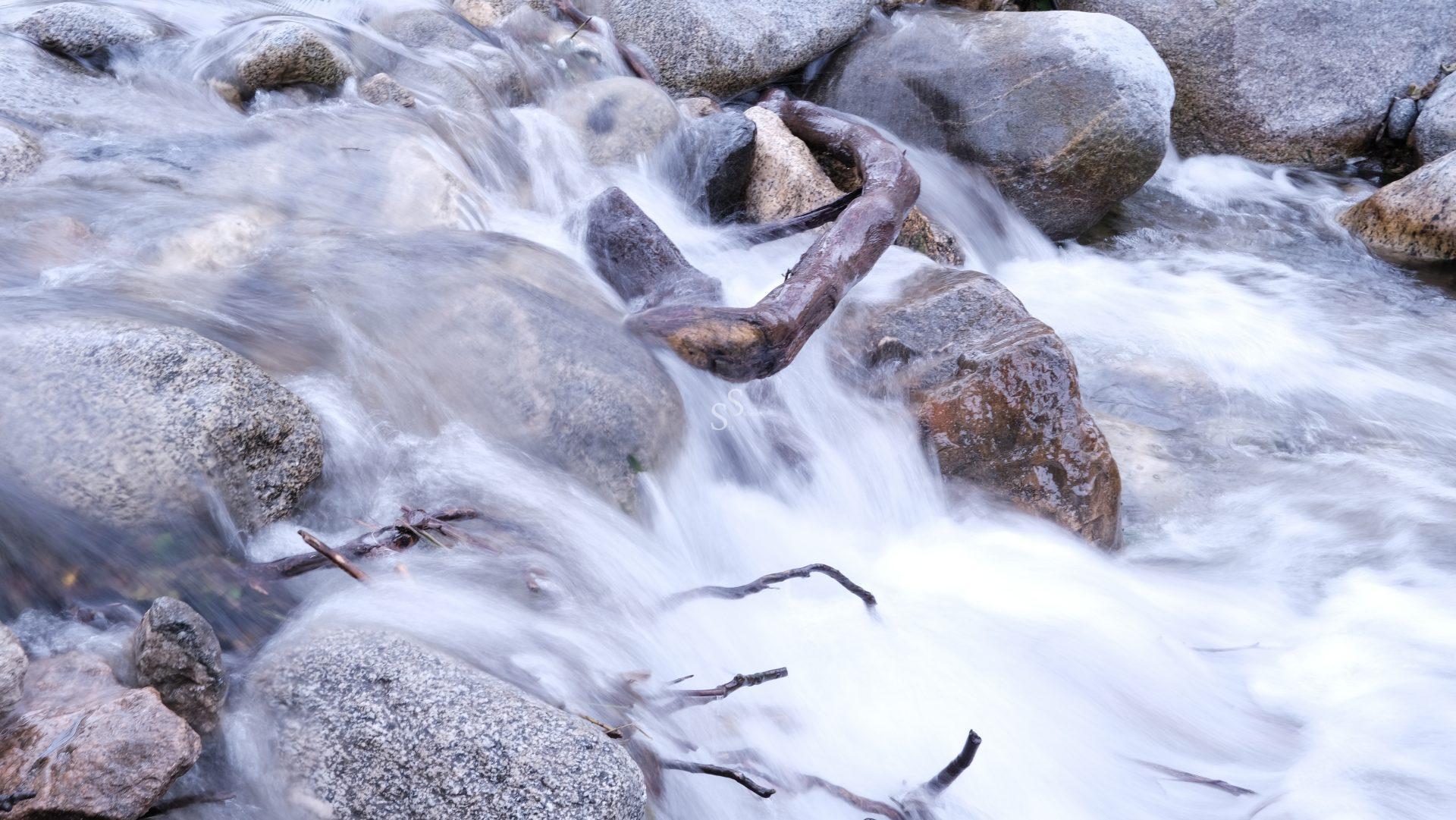 A stream rushing over smooth, large rocks with a twisted piece of driftwood caught among them; the flowing water appears silky due to a long exposure photograph.