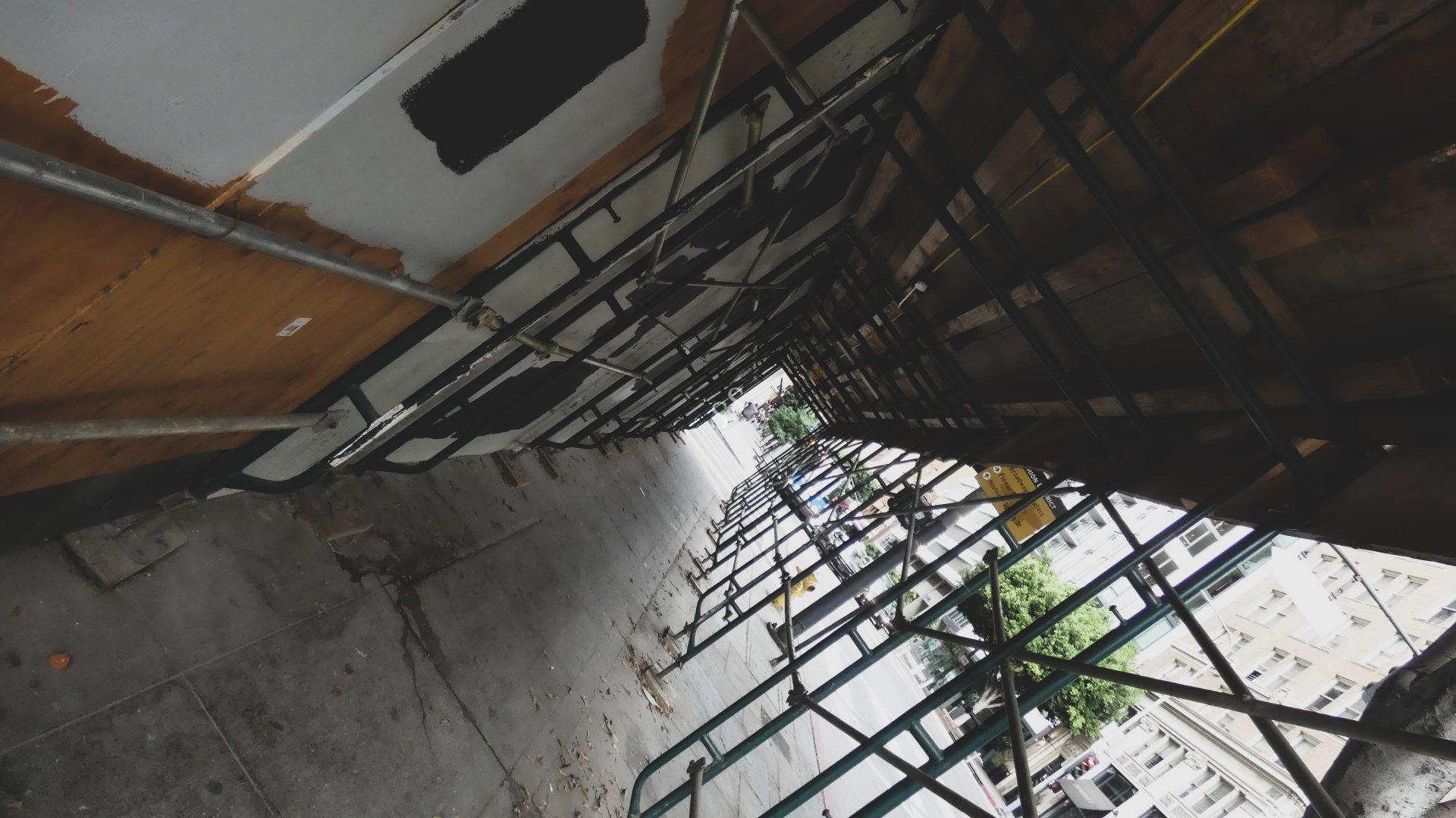 A tilted view of a city sidewalk under scaffolding, with metal bars and wooden planks overhead. Buildings and trees are visible in the background, and the street extends into the distance.
