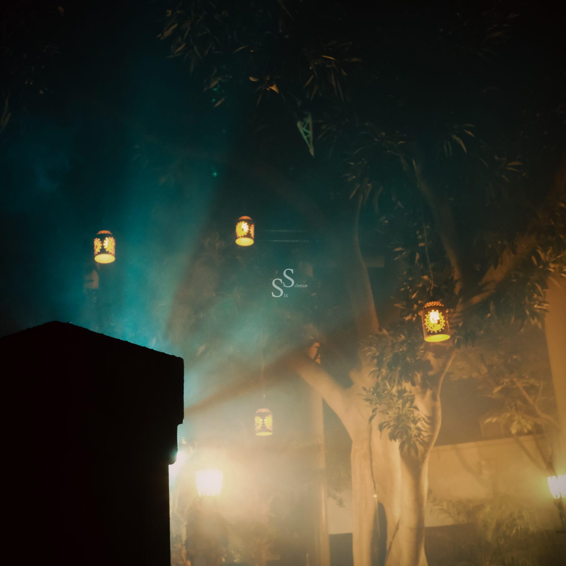 Softly glowing lanterns dangle from tree limbs, piercing the hazy night air. The scene unfolds against a dreamy backdrop illuminated by atmospheric blue and warm yellow lights.