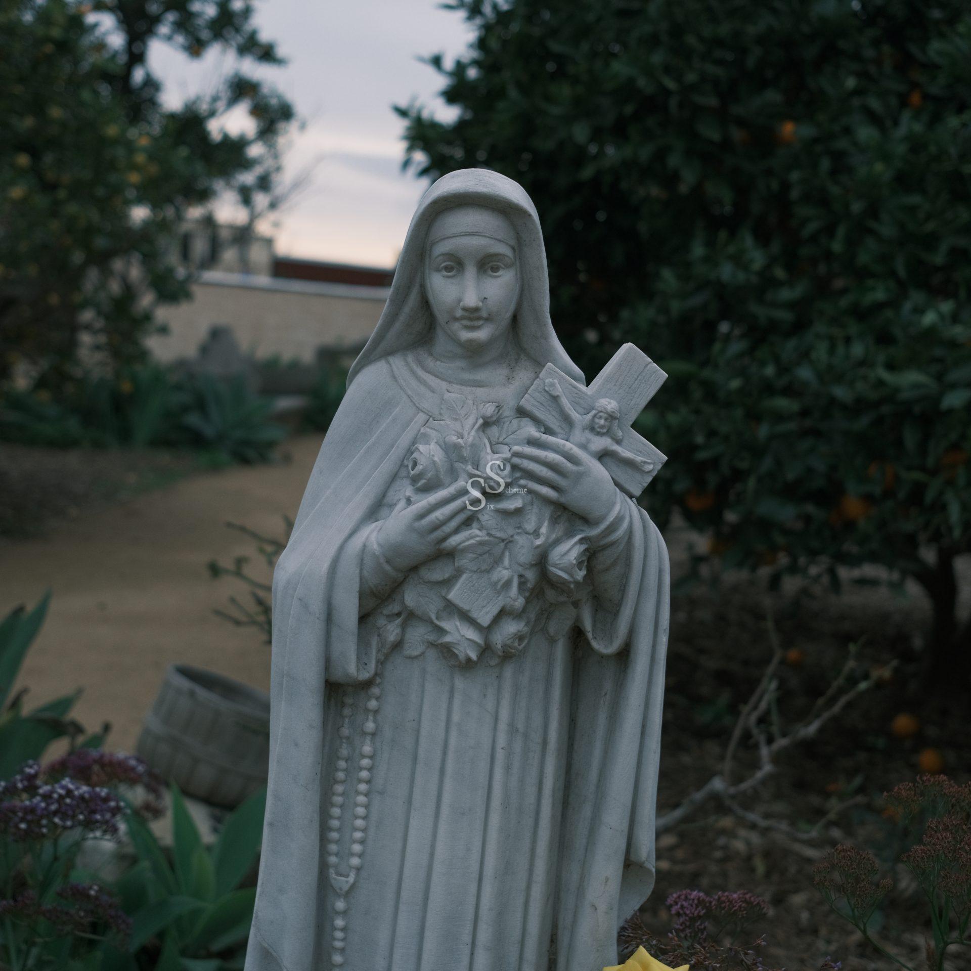 A marble statue of a woman in a robe and veil holding a cross and bouquet of flowers, set in a garden with greenery and trees in the background.