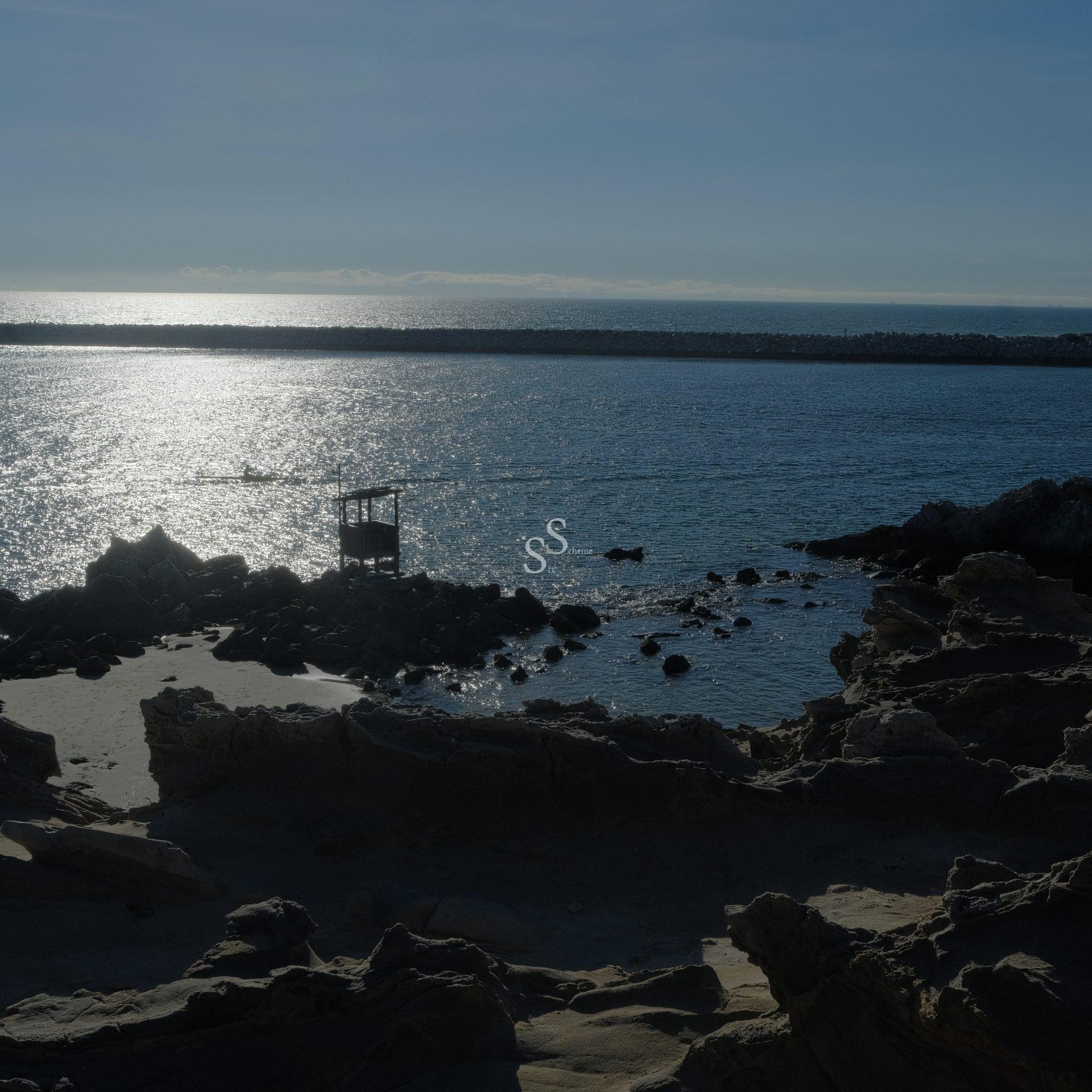 Rocky shoreline at sunset with calm ocean waters, silhouetted rocks, and a small wooden structure extending into the water, all under a clear sky with sunlight reflecting on the sea.