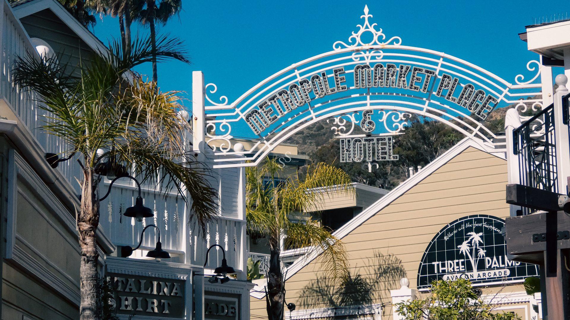 A decorative white metal arch with Metropole Marketplace Hotel stands above a street lined with palm trees and coastal-style buildings, under a clear blue sky.