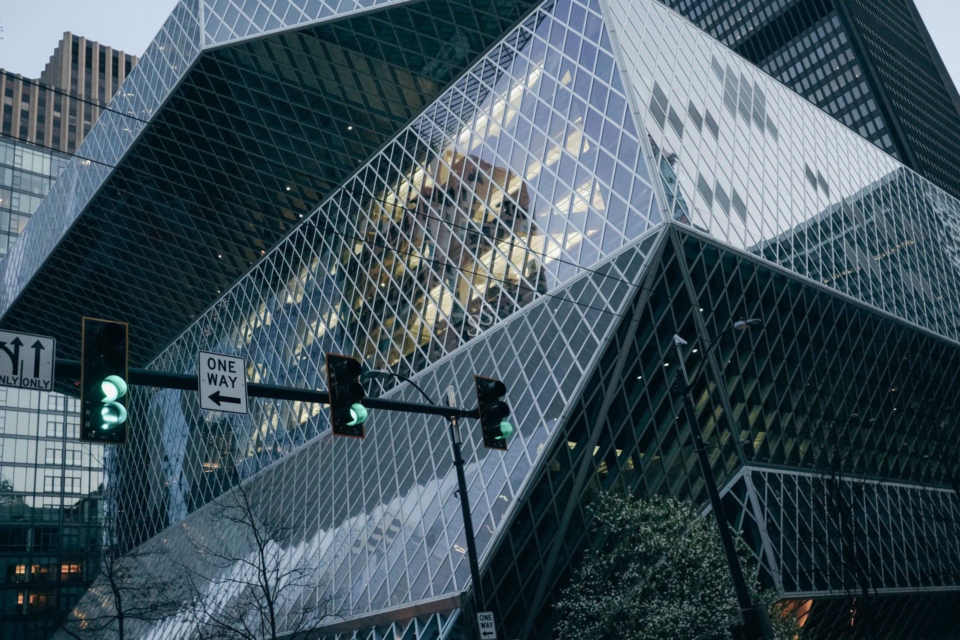 Modern glass building with a crisscross pattern on its facade, reflecting city lights and surrounding skyscrapers; streetlights and a one-way street sign are visible in the foreground.