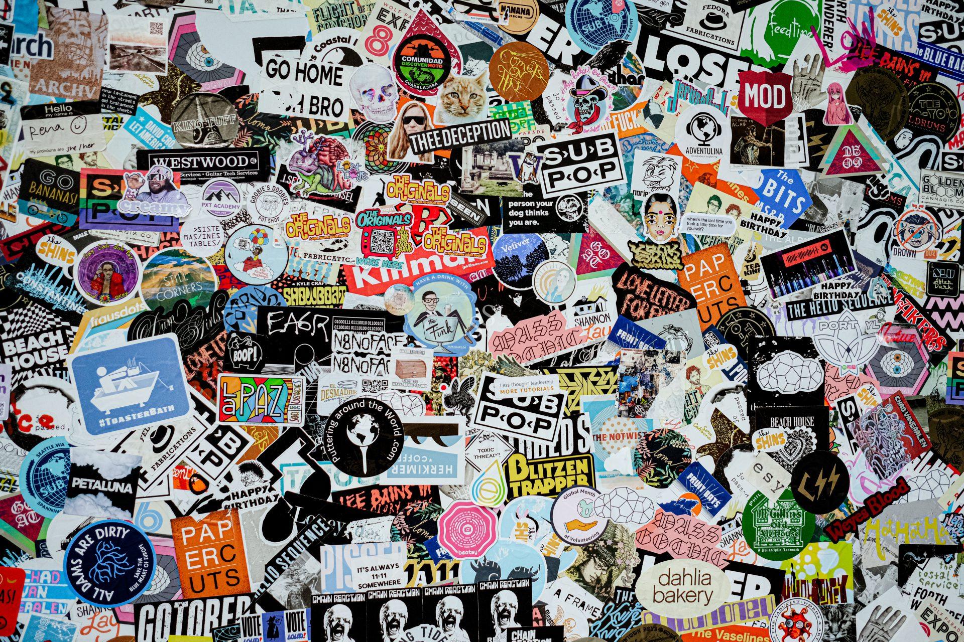 Countless overlapping colorful stickers featuring diverse slogans, logos, drawings, and cartoons densely cover a large wall, forming a vibrant and chaotic visual.