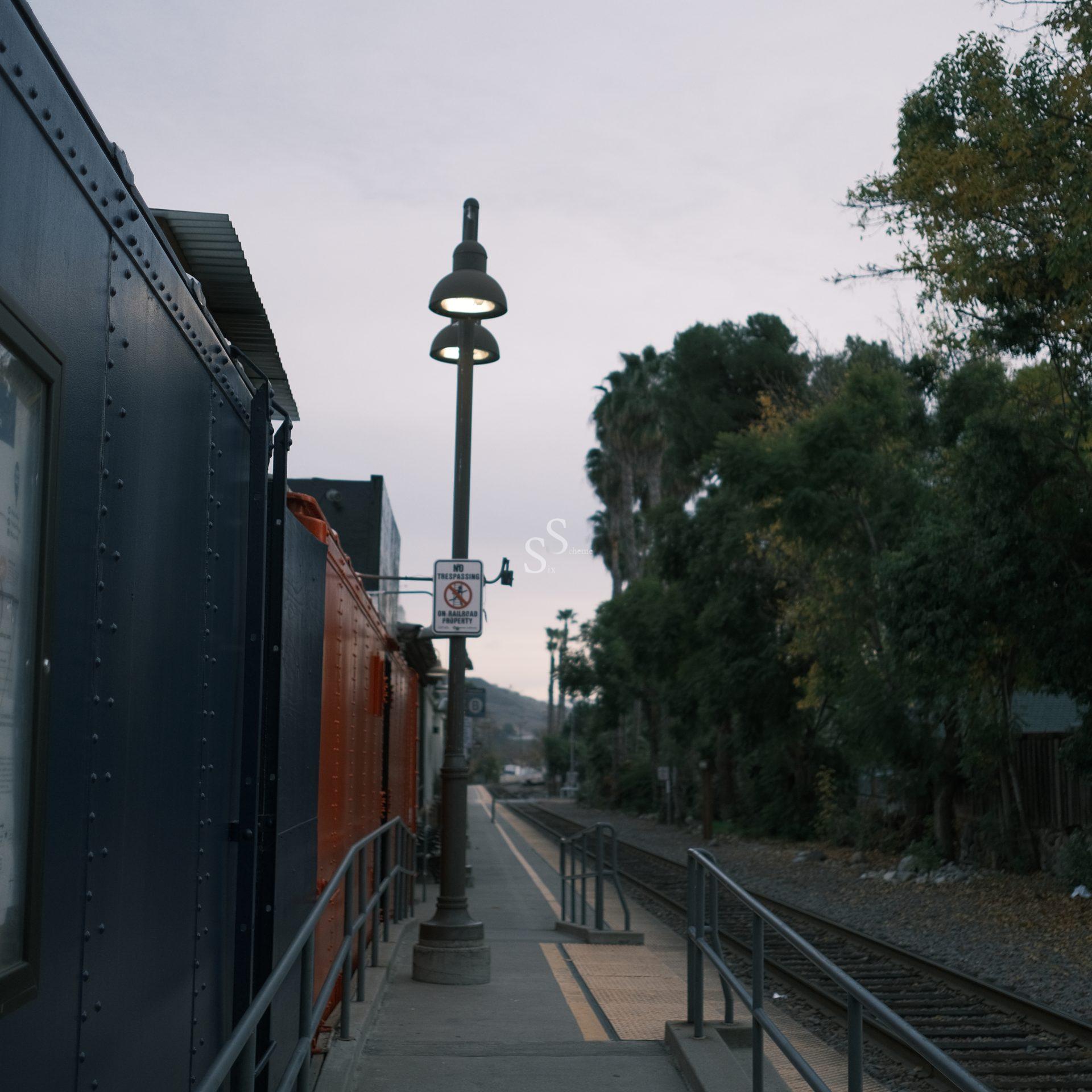 A quiet train station platform with railings, tracks to the right, and a lamppost in the center. Trees line the right side, and a blue and orange train is on the left under an overcast sky.