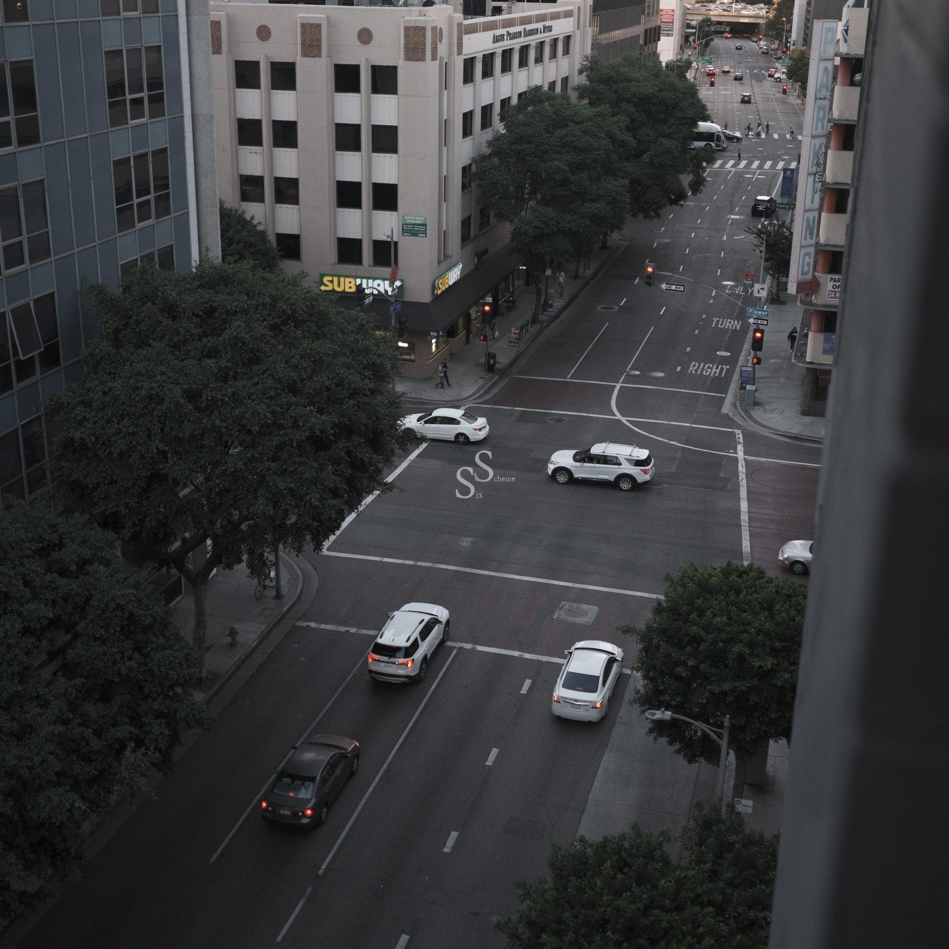 A city street intersection seen from above, with a few cars waiting at the traffic lights. Trees line the sidewalks, and buildings, including a Subway restaurant, border the roads. The scene appears calm and quiet.