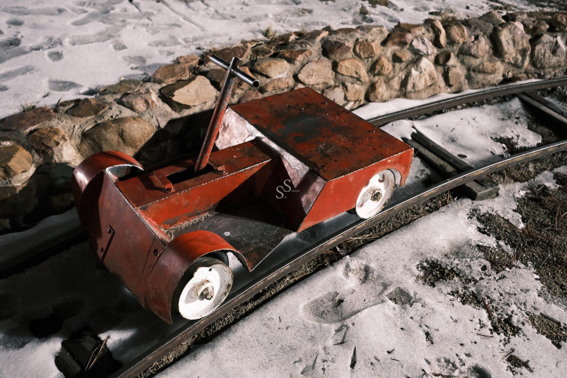 The scene captures a small, red metal handcar on a narrow-gauge track bed composed of snow and gravel. A masonry wall runs parallel to the tracks, lining the edge.