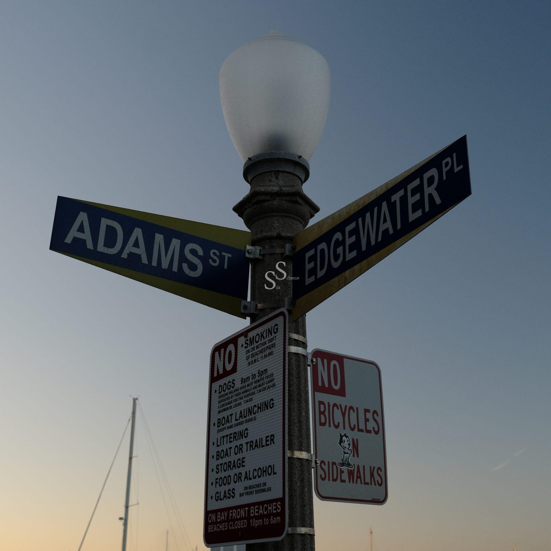 A streetlamp displays street signs for Adams St and Edgewater Pl, along with multiple No signs prohibiting smoking, dogs, and bicycles on sidewalks, against a clear evening sky.