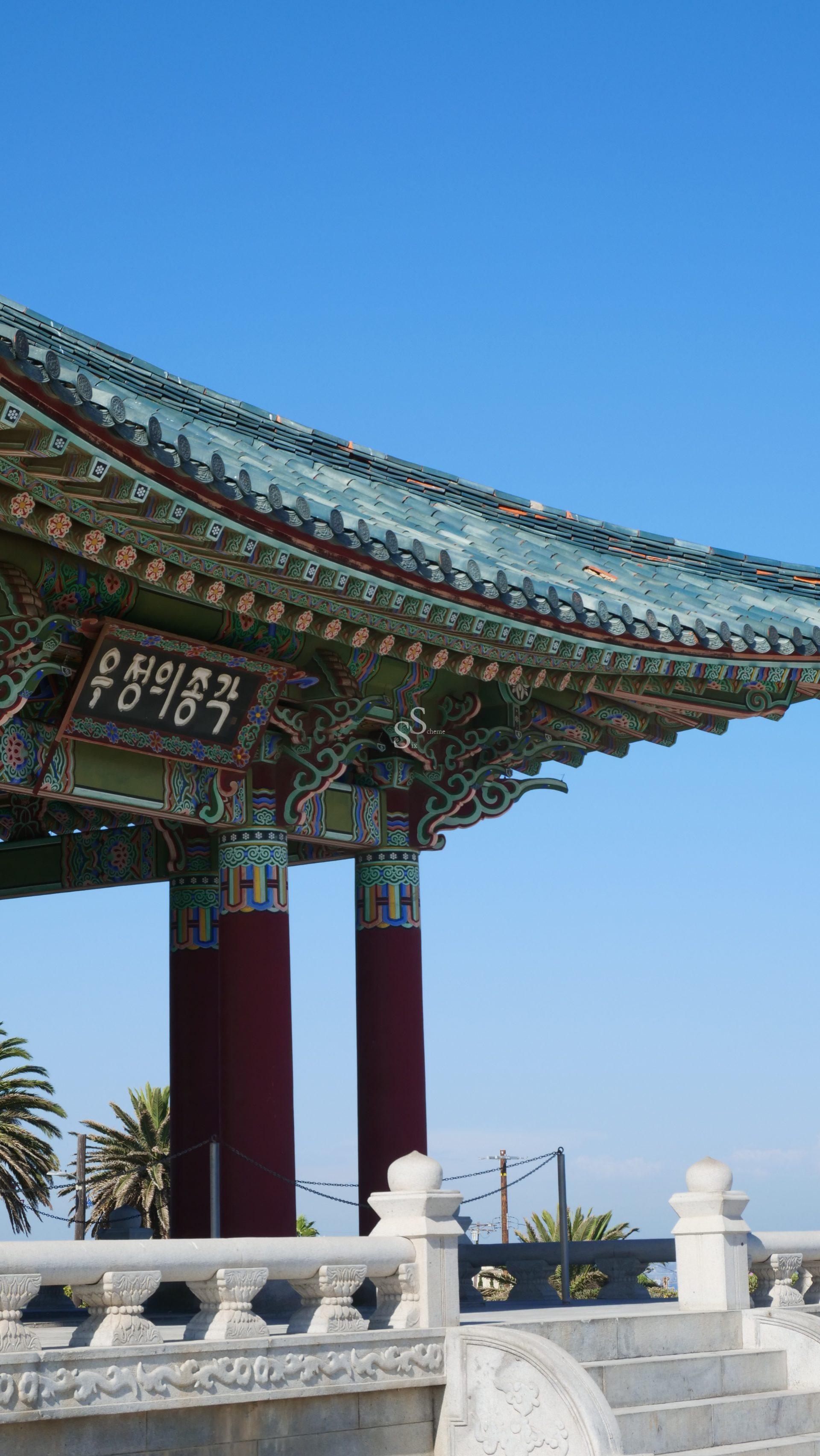 A close-up of a traditional Korean pavilion with ornate, colorful patterns on the roof and columns; palm trees and a clear blue sky are visible in the background.