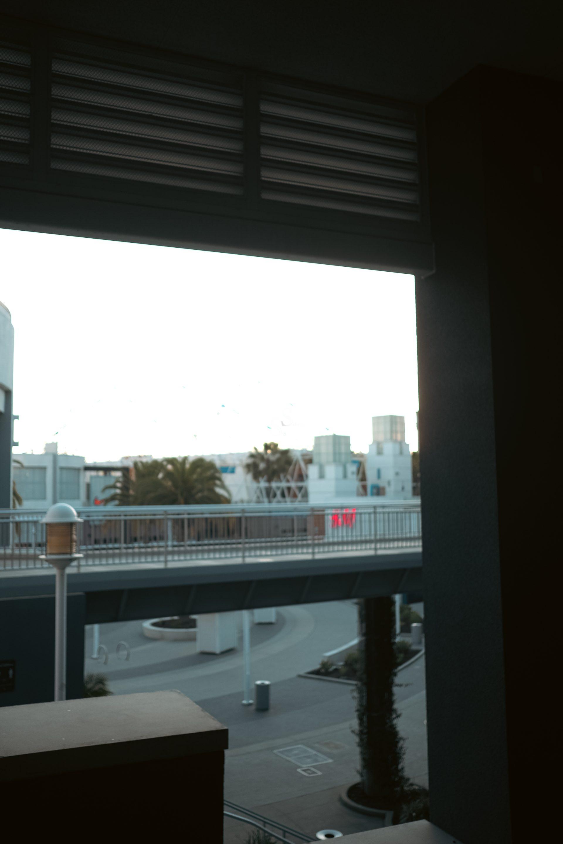 View of an outdoor walkway and buildings with modern architecture, palm trees, and a red digital sign, seen through a shaded balcony. The lighting is dim in the foreground and bright outside.
