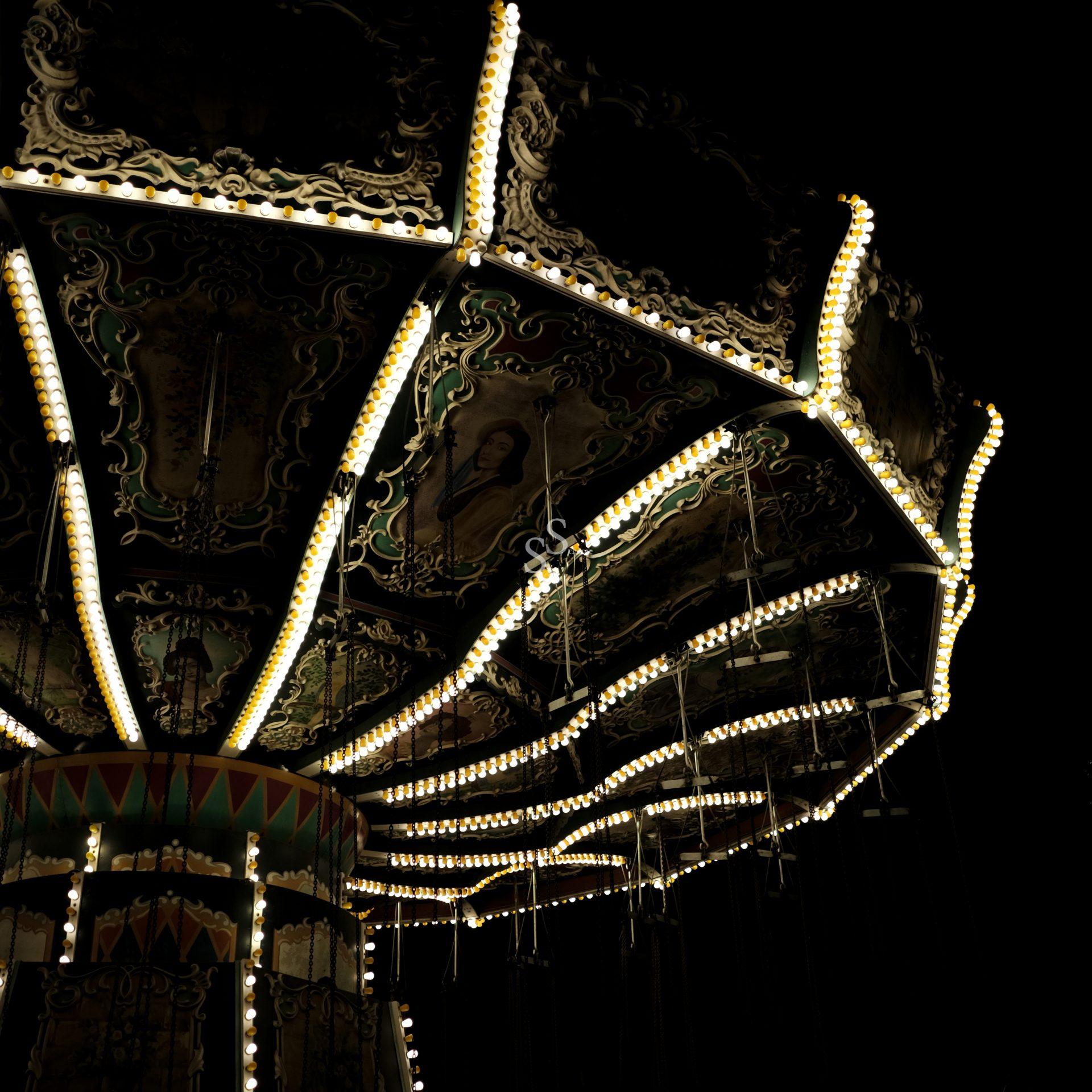 Illuminated brightly against the night, a carousel's ornate top and glowing rows of lights dominate the view, with the background barely visible.