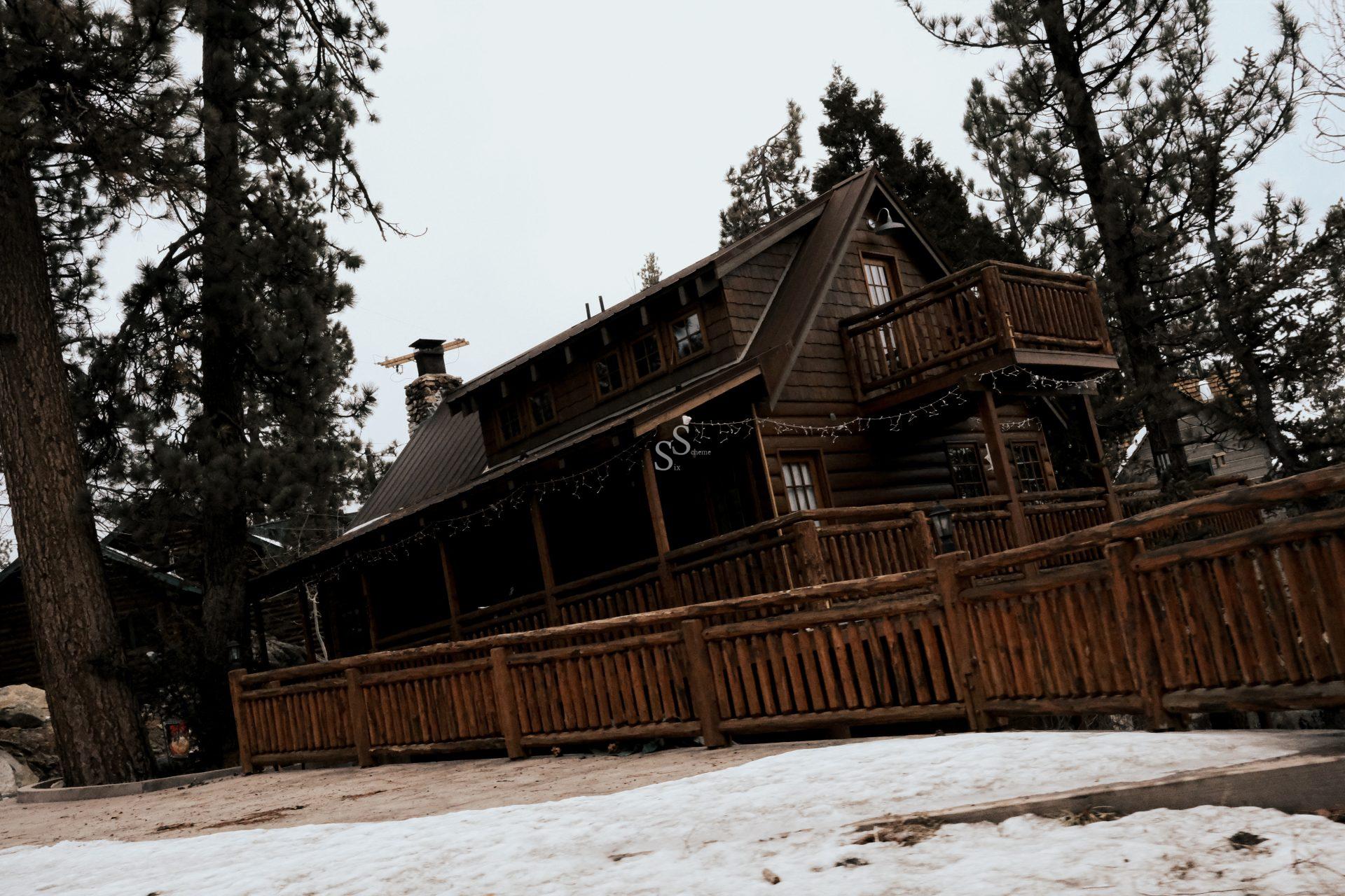 A brown wooden cabin with balconies and a large porch sits among tall pine trees, surrounded by a wooden fence. Snow covers the ground, and the sky is overcast.