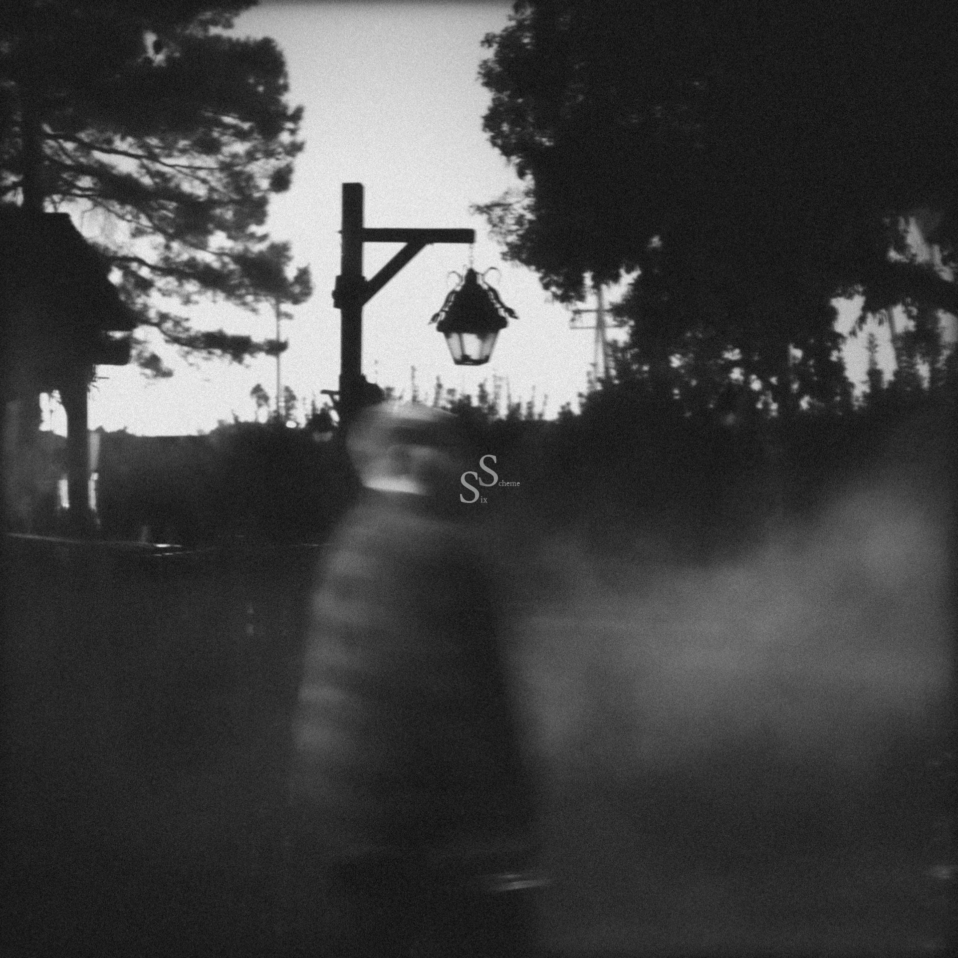 A blurred figure walks through a foggy outdoor scene at dusk, with a lantern hanging from a wooden post and dark trees silhouetted against the sky in the background. The image is in black and white.
