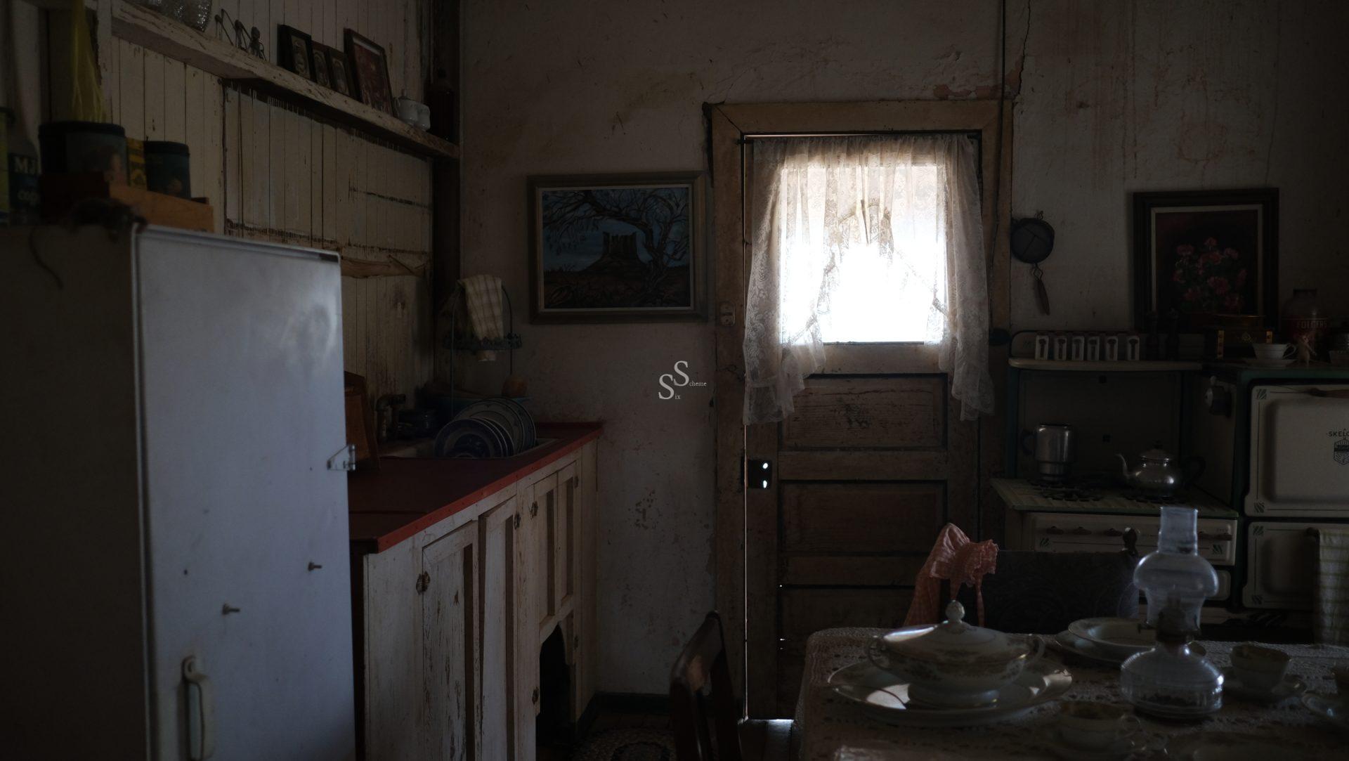 A dimly lit, rustic kitchen with old cabinets, a fridge, lace curtains on the door, vintage kitchenware on the table, and paintings on the worn, textured walls. Sunlight filters softly through the window.