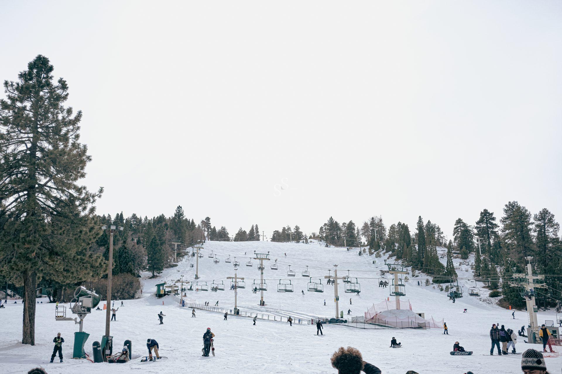 A snowy ski resort with people skiing and snowboarding, ski lifts ascending the slope, and pine trees surrounding the area under an overcast sky.